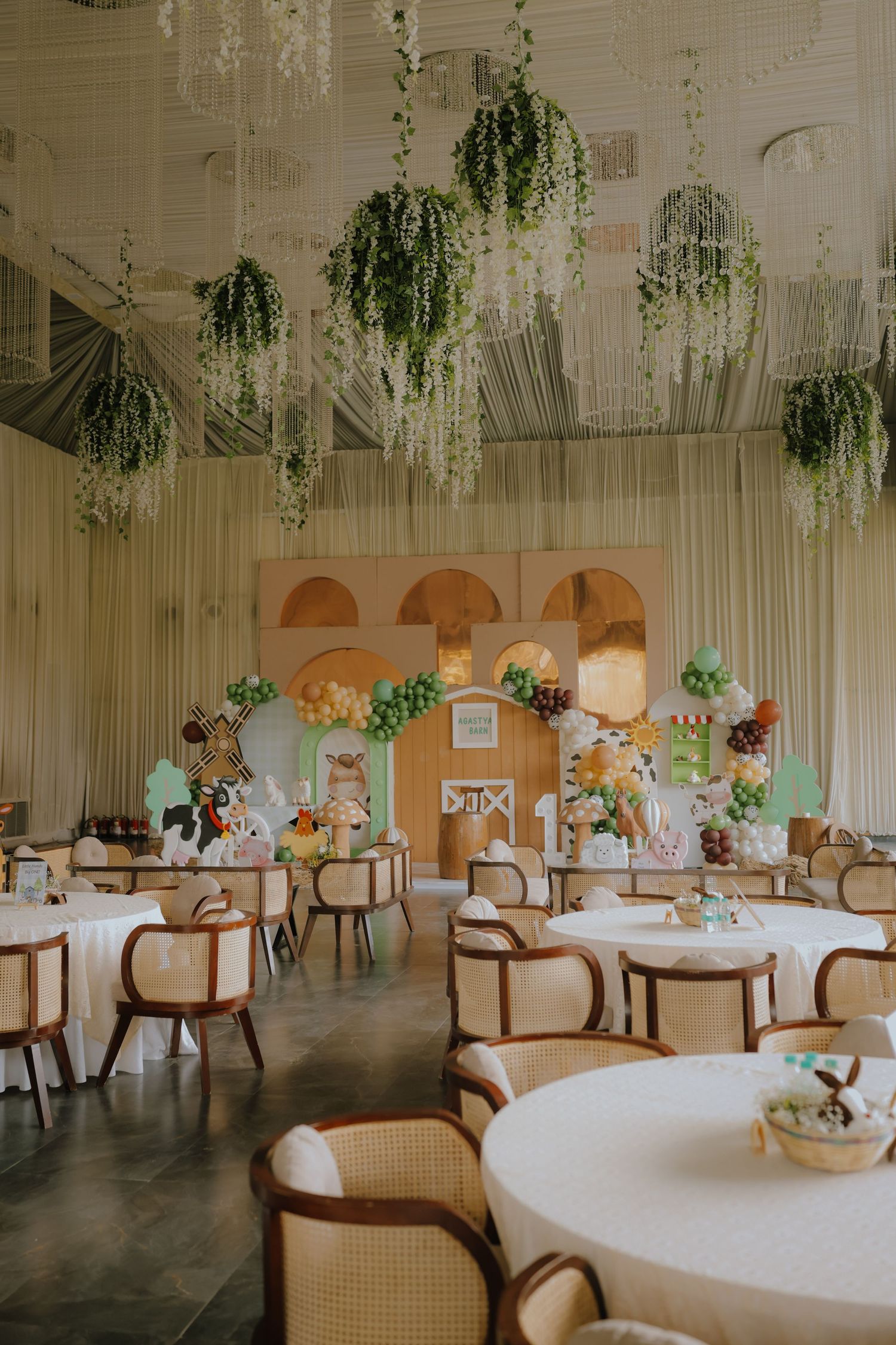 Elegant event hall with hanging greenery, round tables, wicker chairs, and a balloon arch backdrop for a garden-themed party.