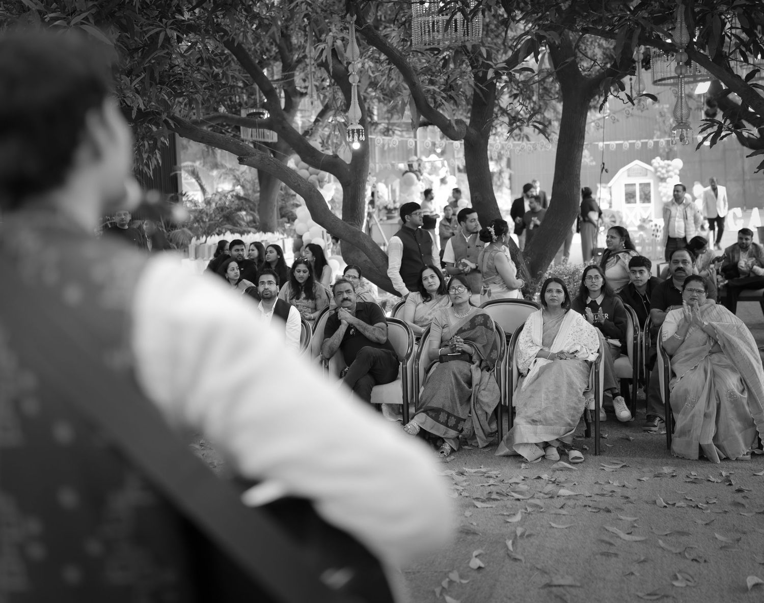 Black and white photo of a crowd gathered outdoors under trees, watching a speaker at an Indian cultural event.