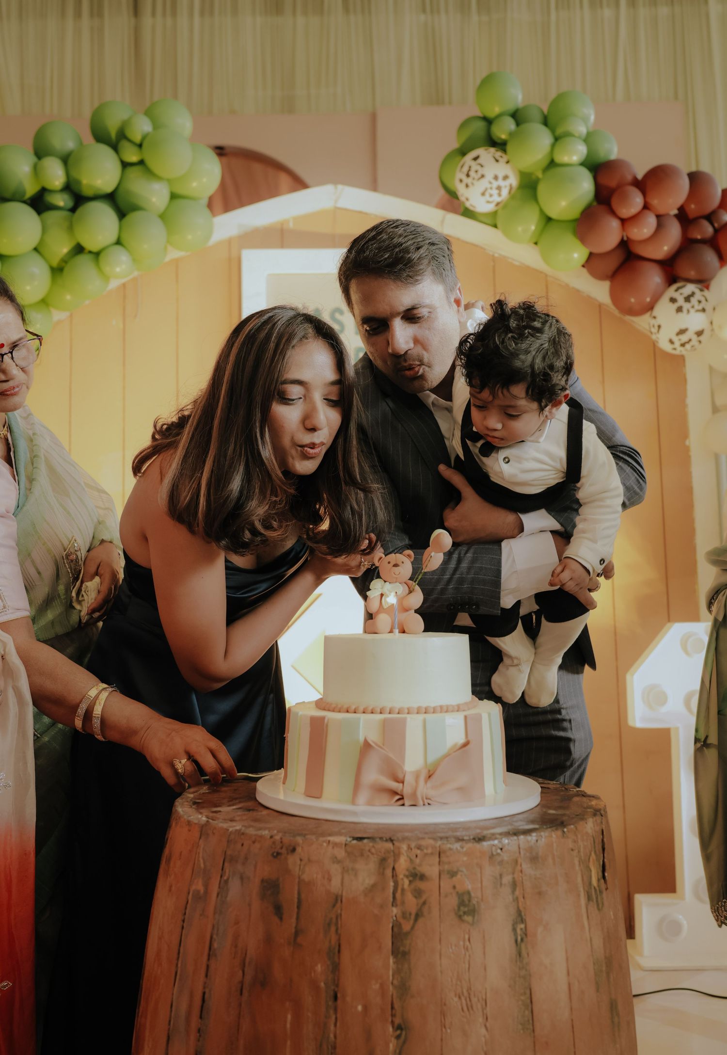 Family celebrating baby’s first birthday with a two-tier cake adorned with a bear topper at a festive party.