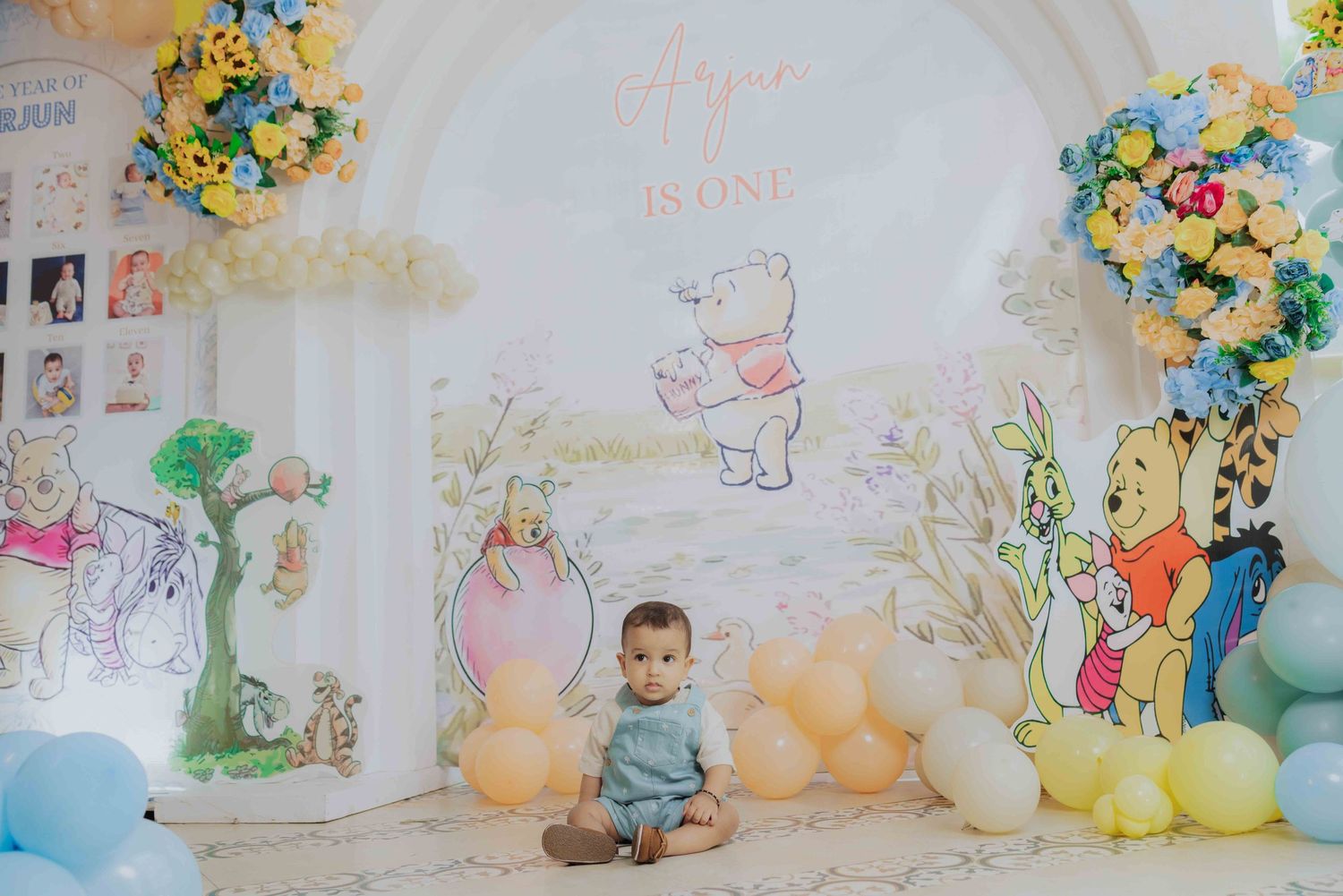 Baby sitting in front of a Winnie the Pooh first birthday backdrop with colorful balloons and floral arrangements.