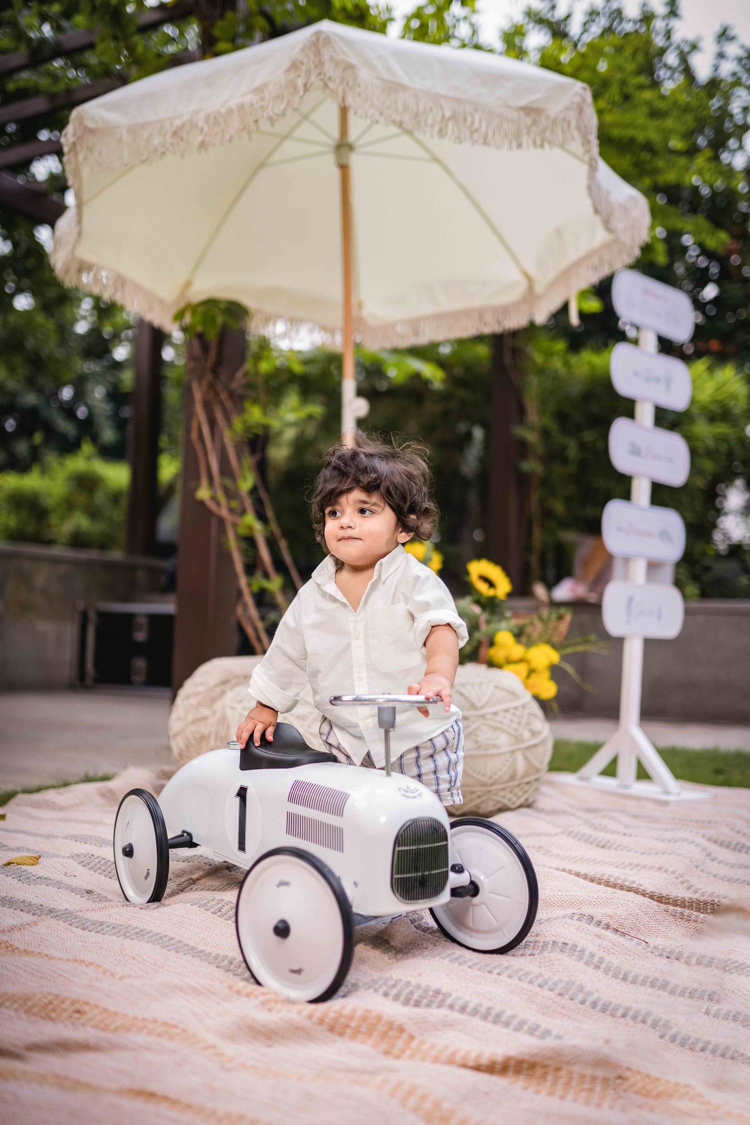 Toddler boy sitting on a white vintage toy car under a cream fringed umbrella at an outdoor garden party.