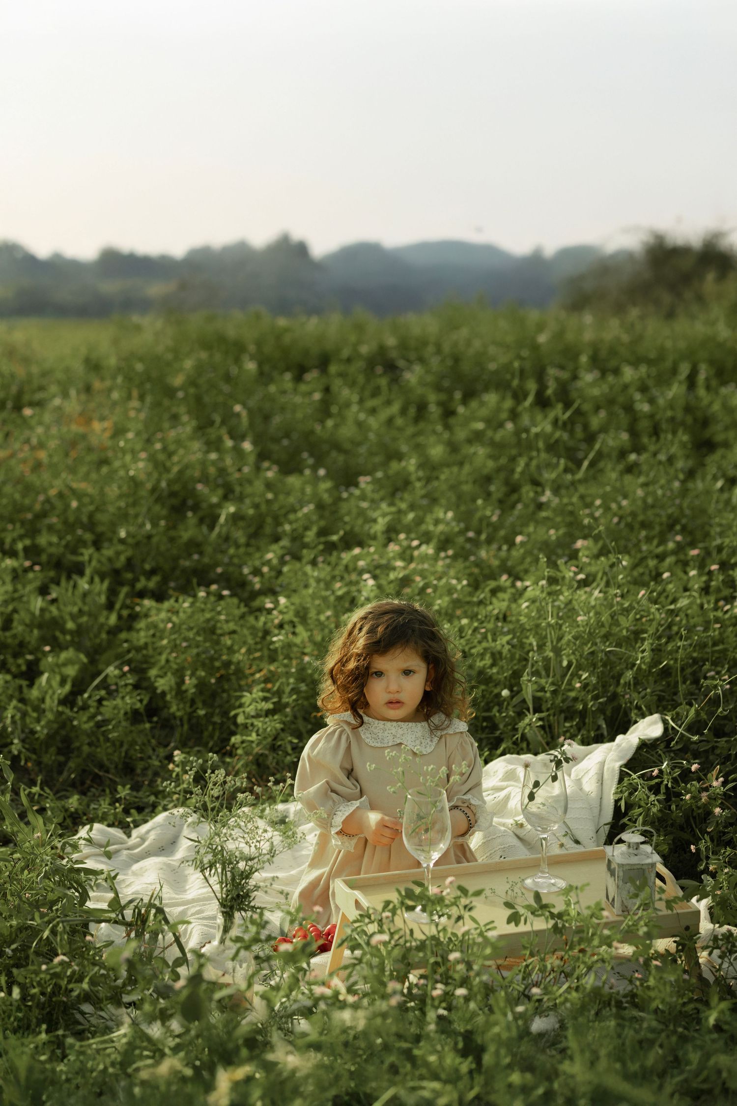 Young girl in white dress sitting on blanket in lush green field for an outdoor picnic.