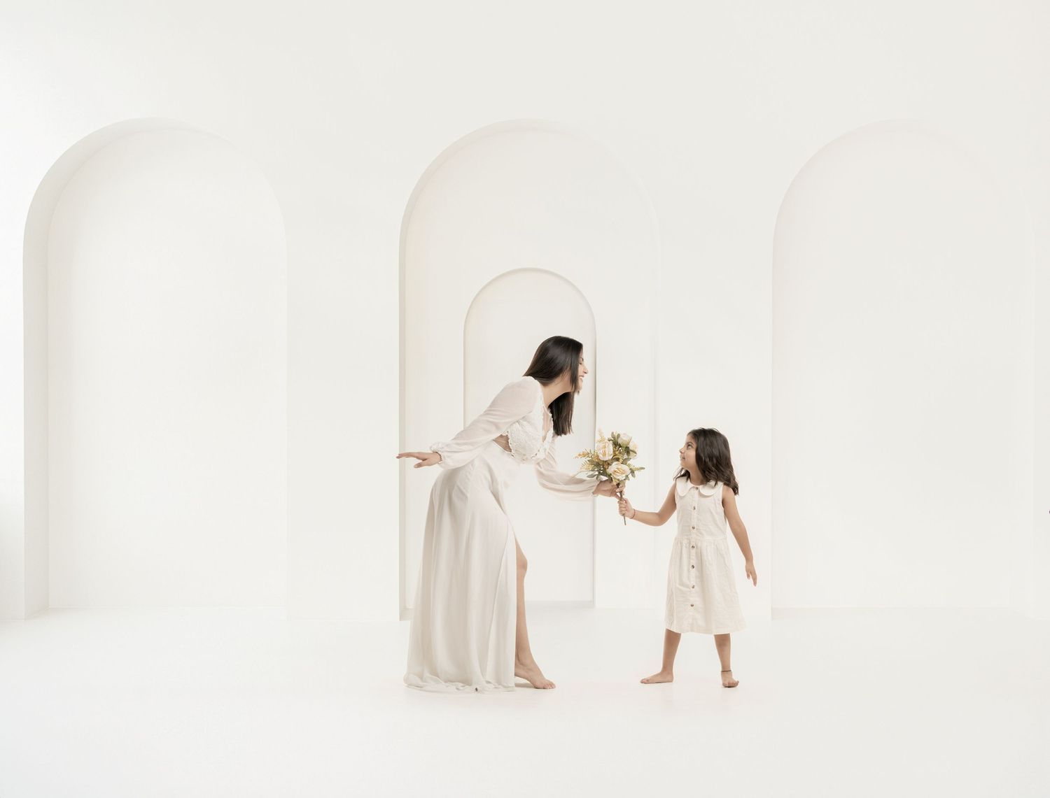 Bride in white gown hands flowers to young girl in white dress amid elegant arched white hallway.