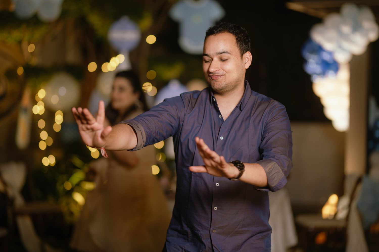 Man in gray shirt dancing with outstretched hands at an indoor party with bokeh lights in background.