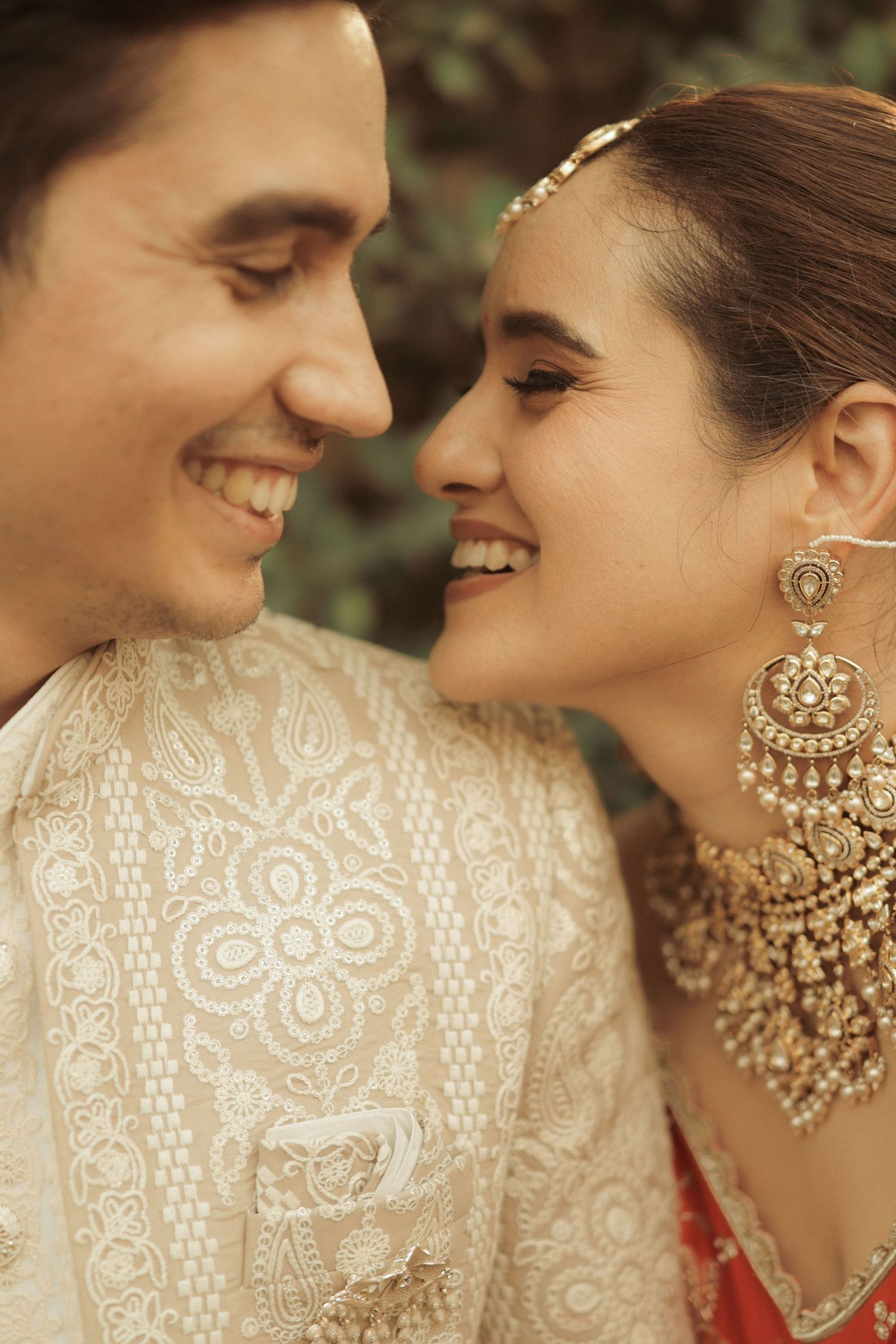 Smiling couple in traditional Indian wedding attire, bride wearing gold chandbali earrings and necklace, groom in embroidered sherwani.