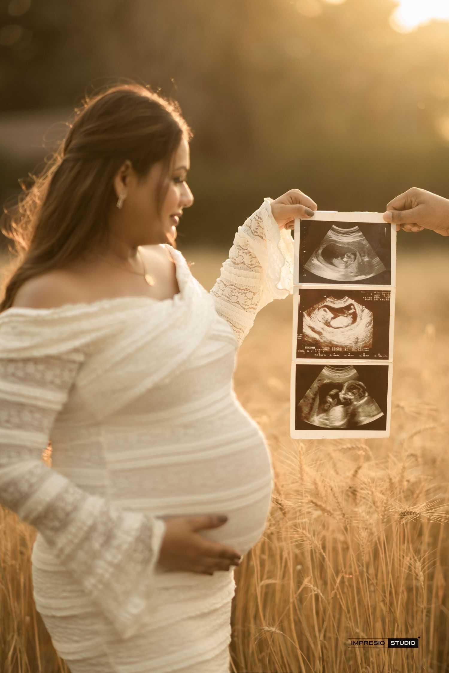 Pregnant woman in white dress holding ultrasound photos in golden wheat field during maternity photoshoot at sunset.