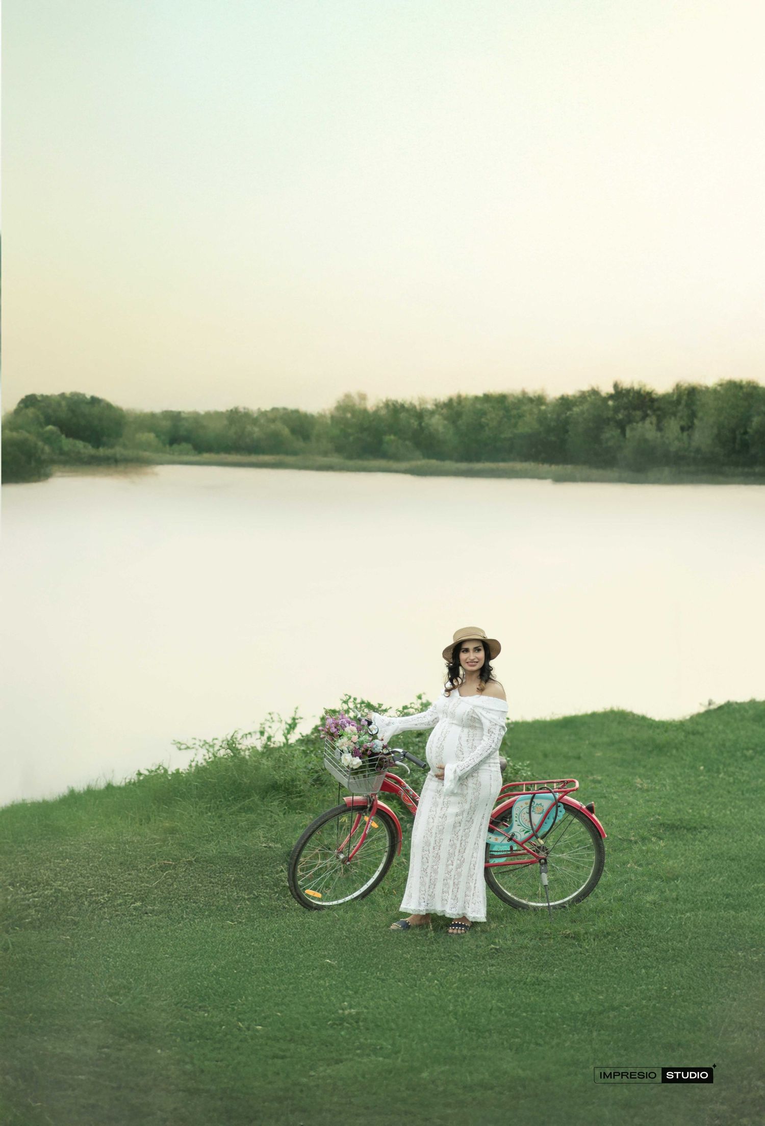 Pregnant woman in white lace dress and straw hat stands by red bicycle near serene lake at sunset.