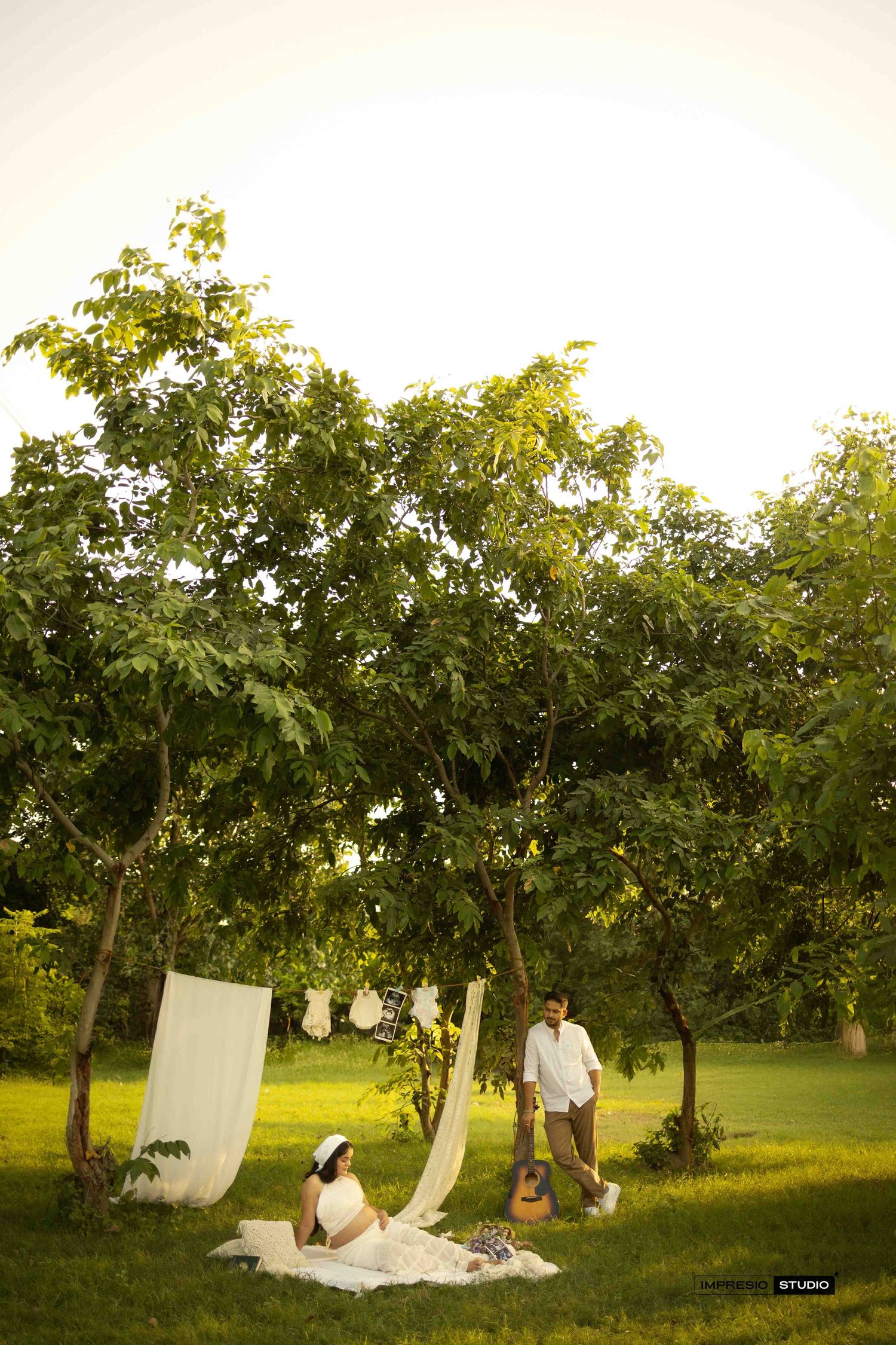 Couple in white outfits relaxing under lush green trees with draped fabric and guitar in a serene garden setting.