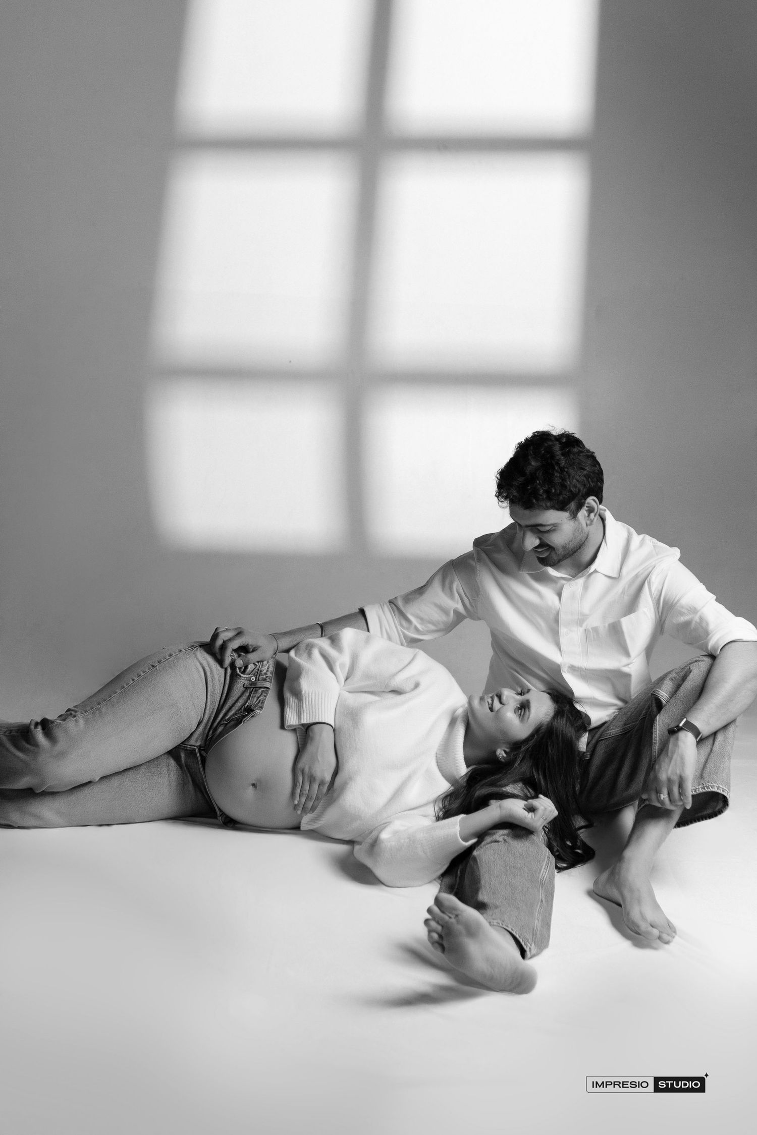 Black and white maternity photo of a pregnant woman lying down with her partner in a studio setting.