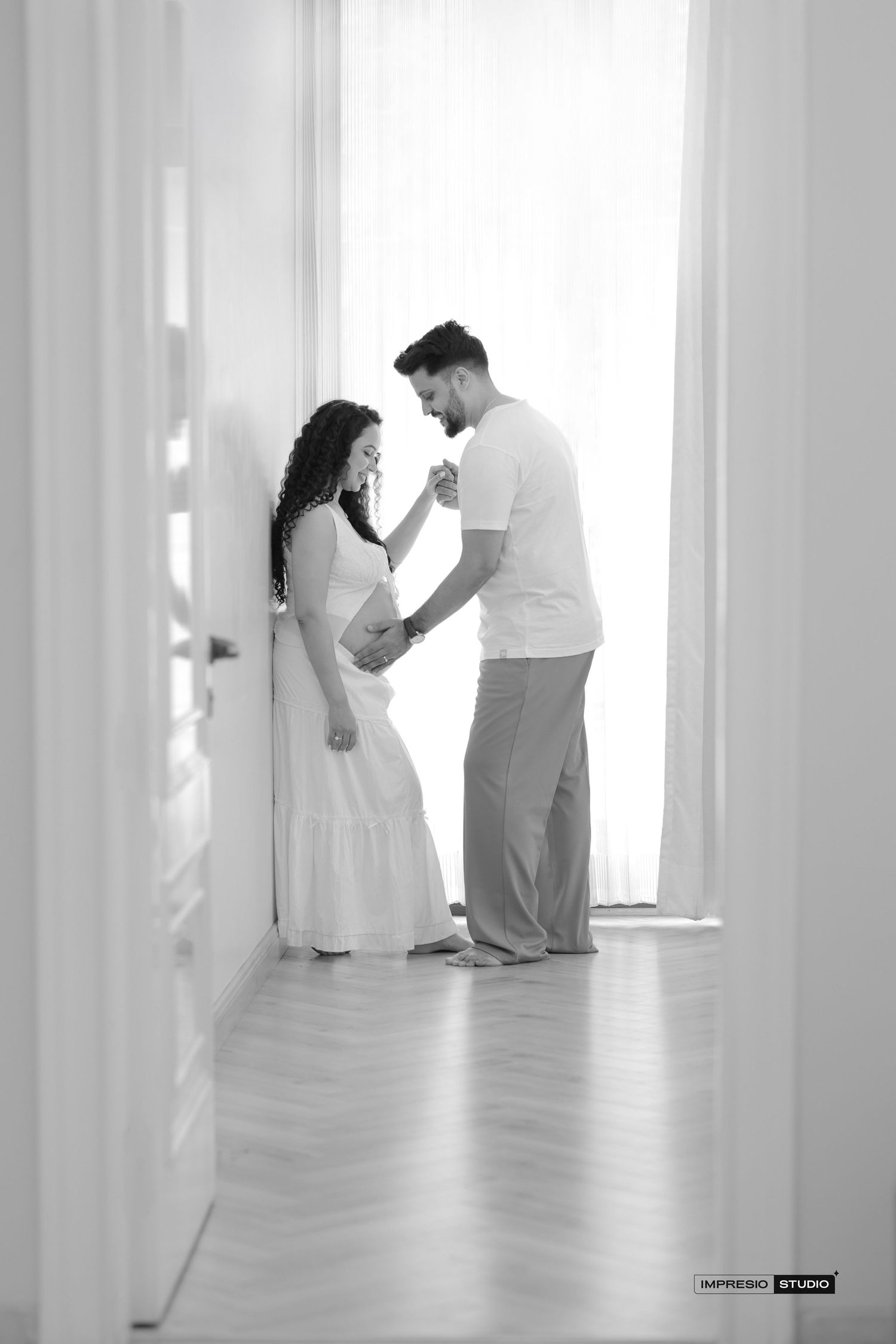 Black and white maternity photo of a couple tenderly holding hands in a sunlit hallway, woman in white dress.
