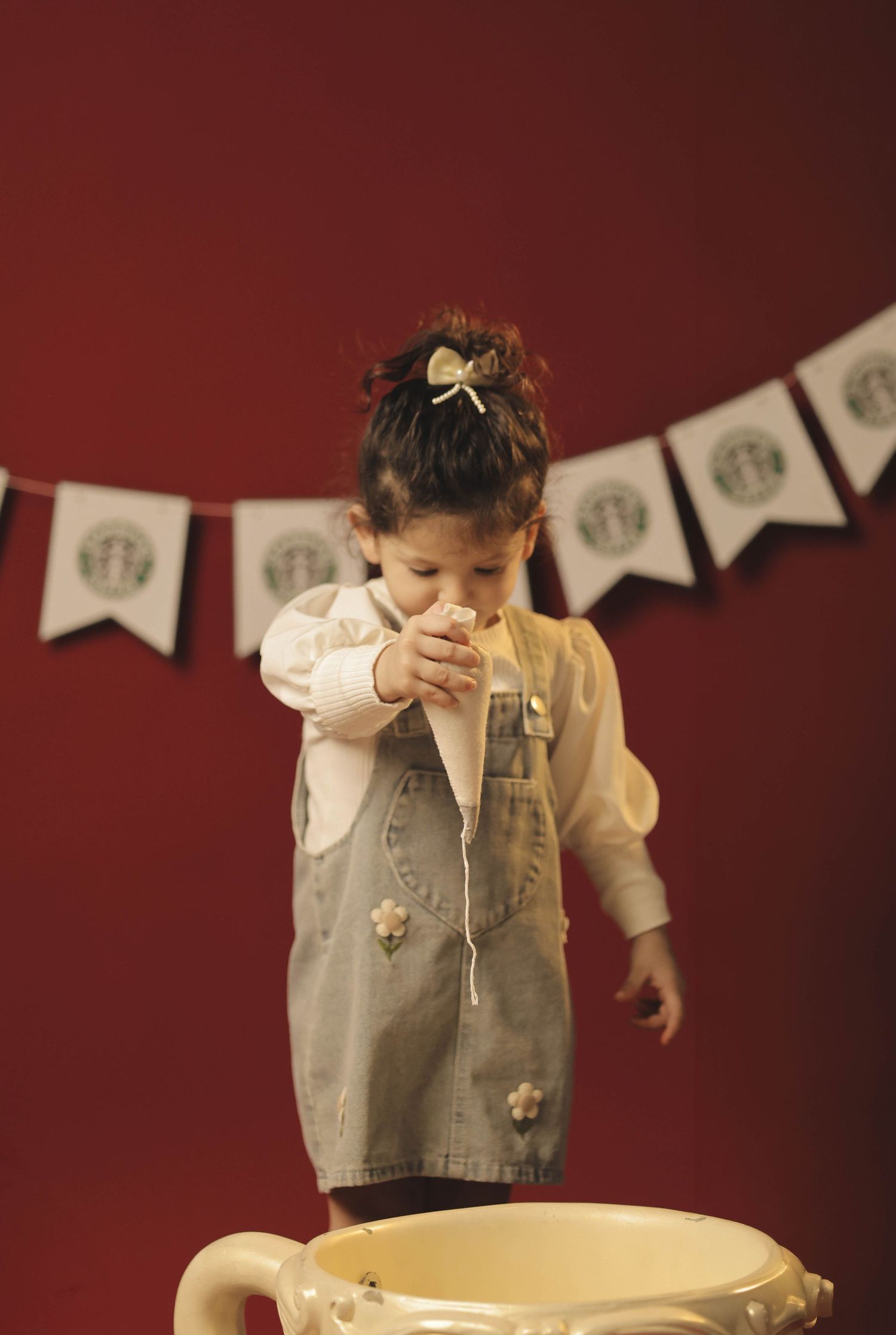 Toddler girl in denim vest standing over large teacup with Starbucks banner on red wall.