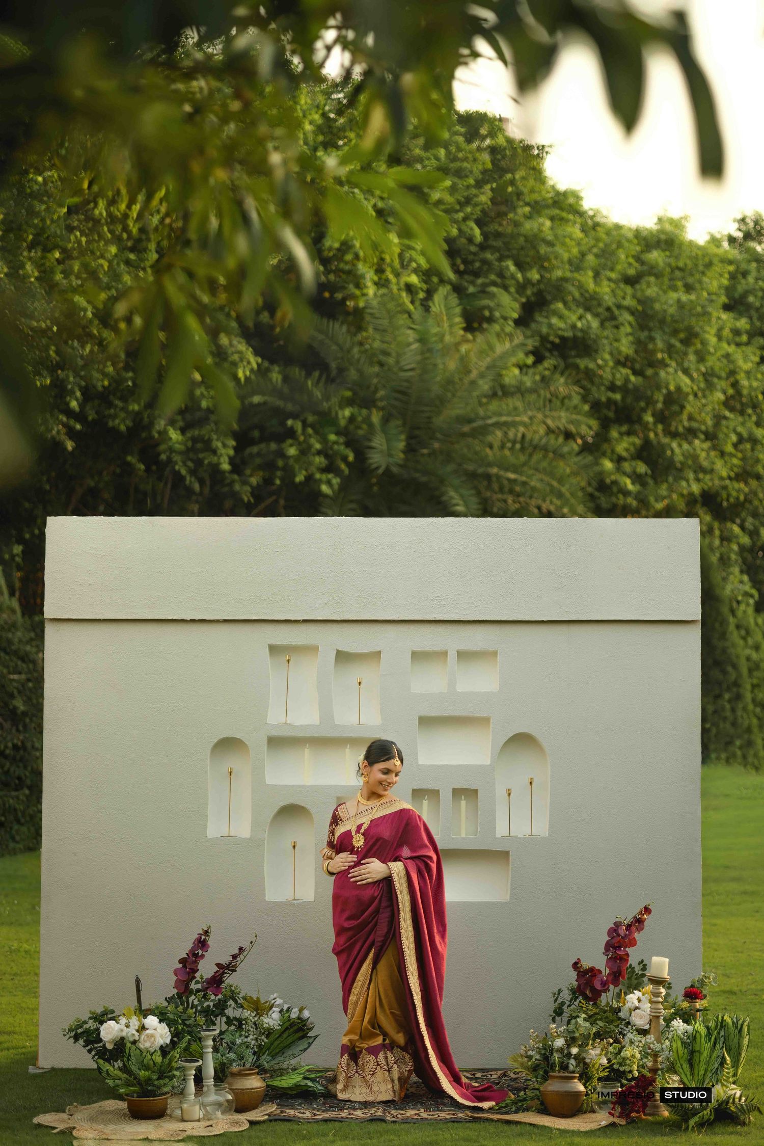 Woman in maroon and gold saree posing before ornate white backdrop with floral decor in lush garden setting.