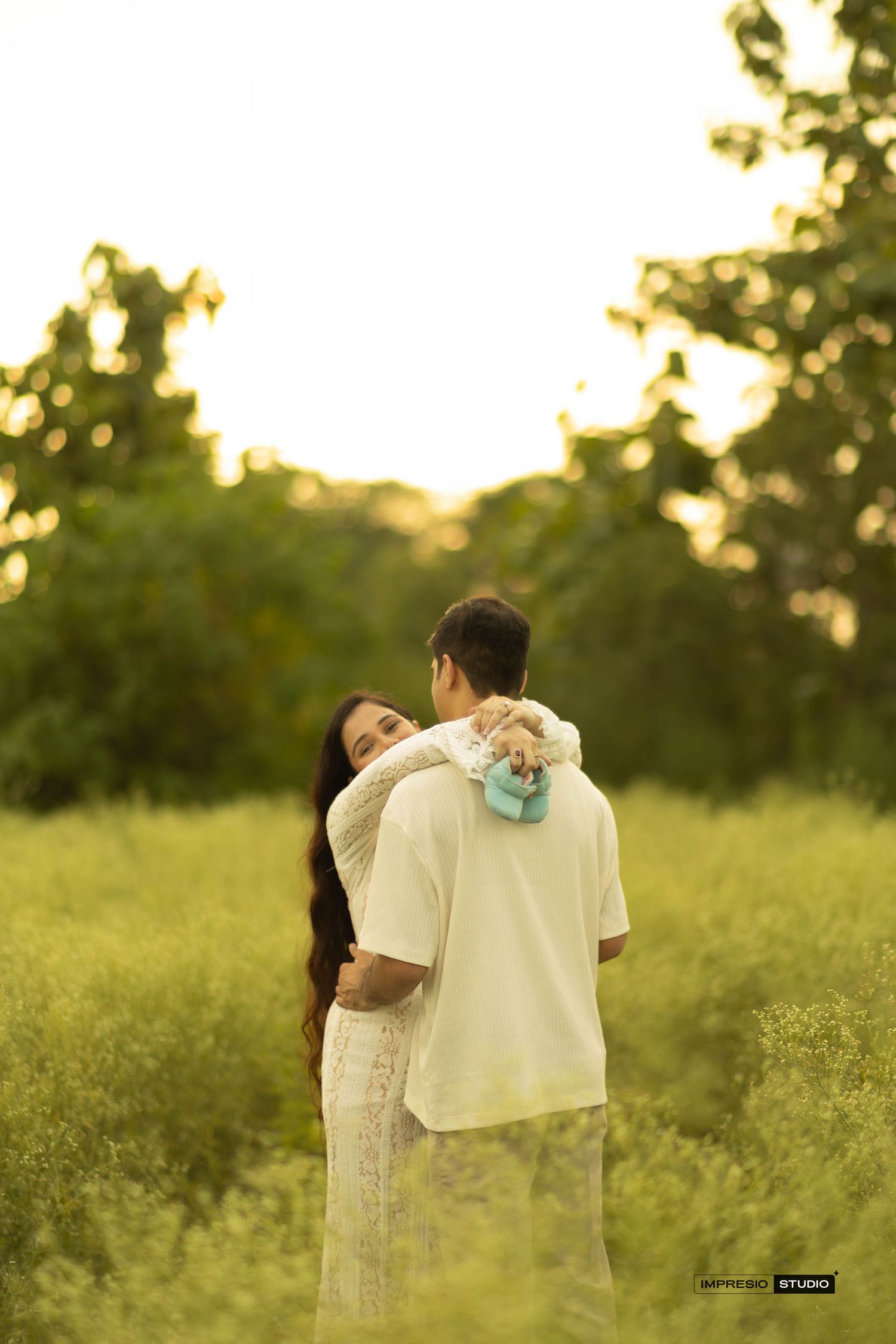 A couple embraces tenderly in a lush green field at golden hour, the woman resting her head on the man's shoulder.