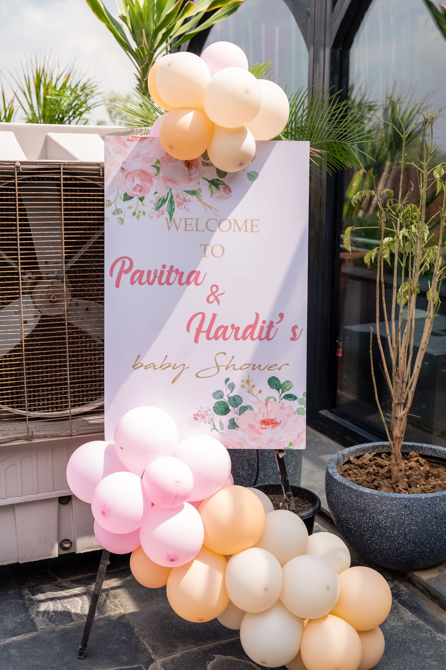 Pink and peach balloon arch decorating a welcome sign for Pavitra and Hardit's baby shower celebration outdoors.