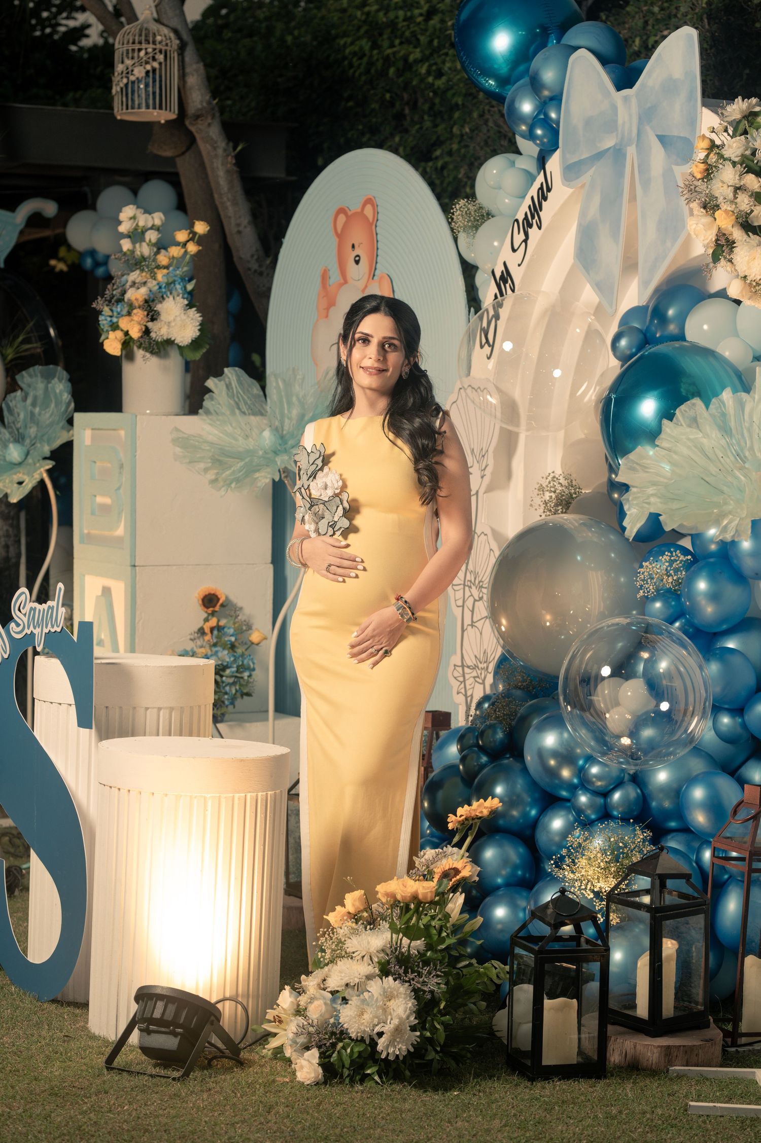 Pregnant woman in yellow dress poses at blue balloon baby shower decoration with bear theme and floral arrangements.
