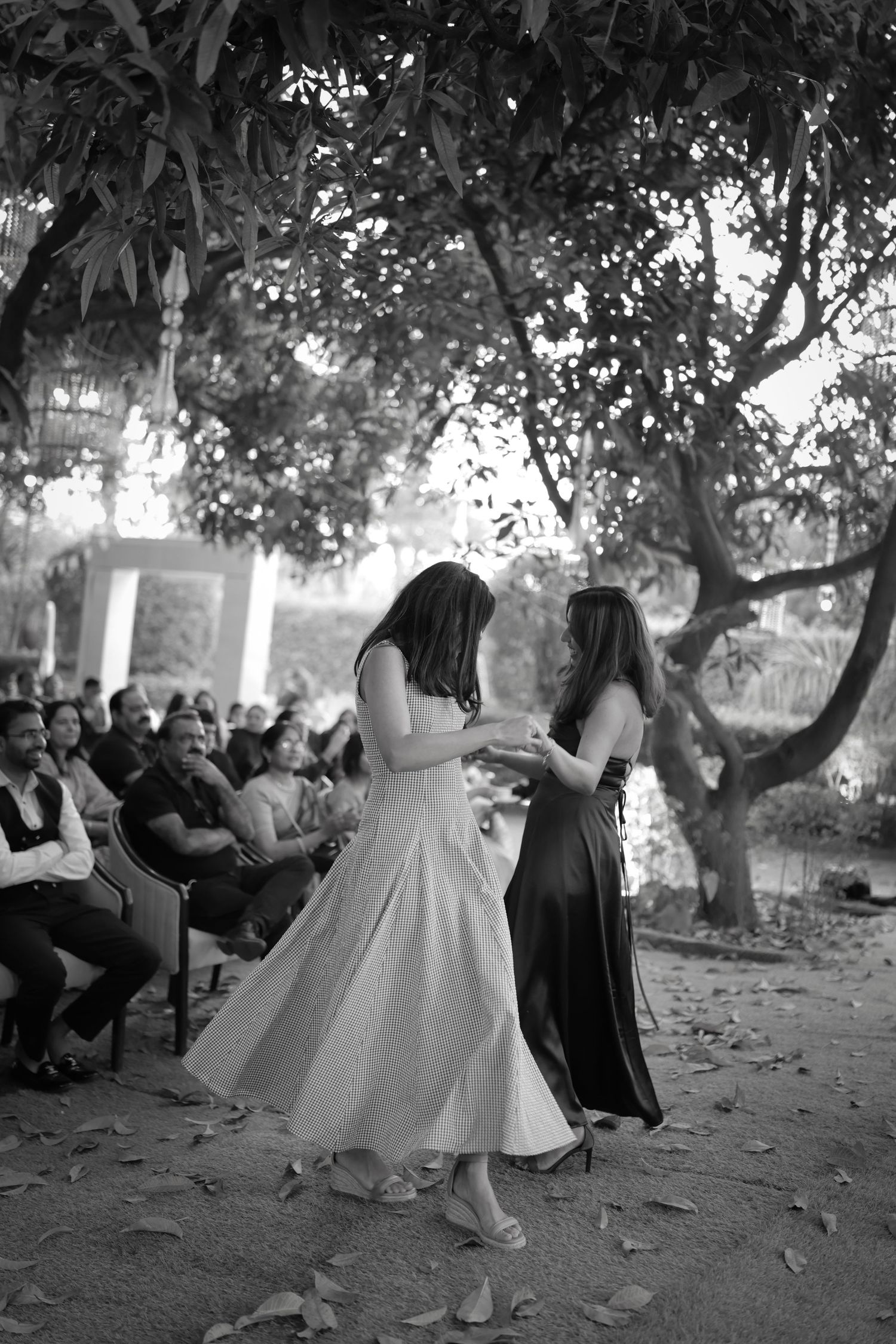 Two women dance together outdoors at a wedding ceremony, surrounded by guests and trees, in black and white.