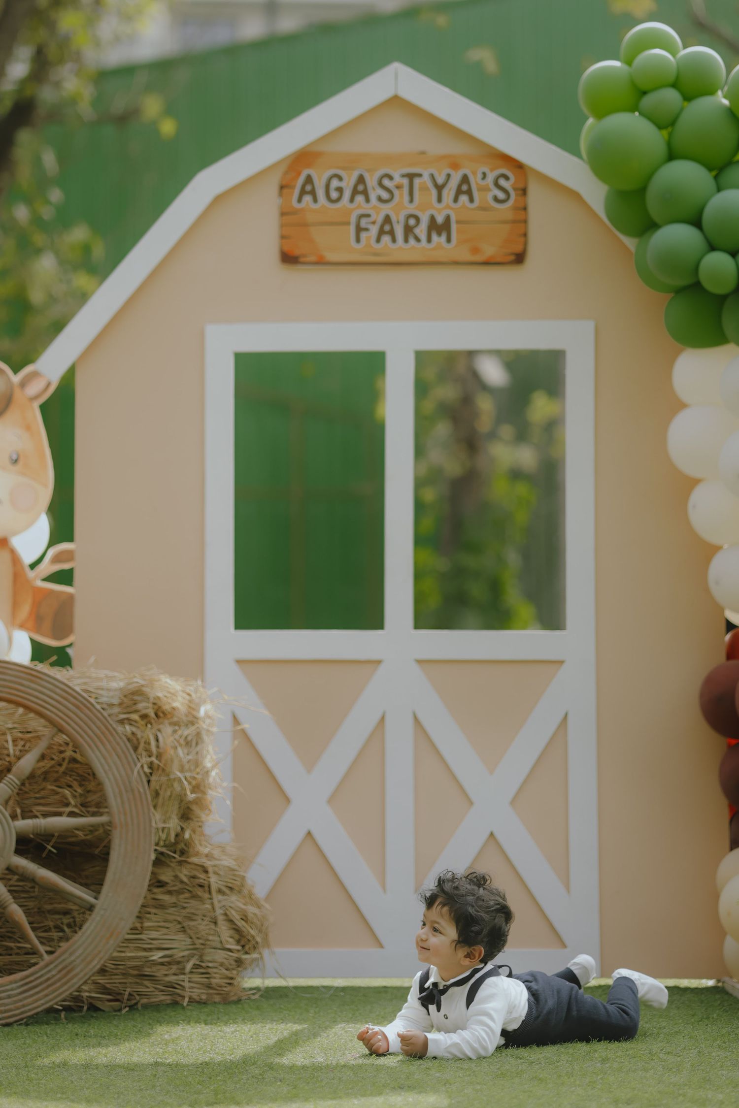 Baby boy lying on grass in front of Agastya's Farm barn backdrop with green balloons and hay bales at birthday party.