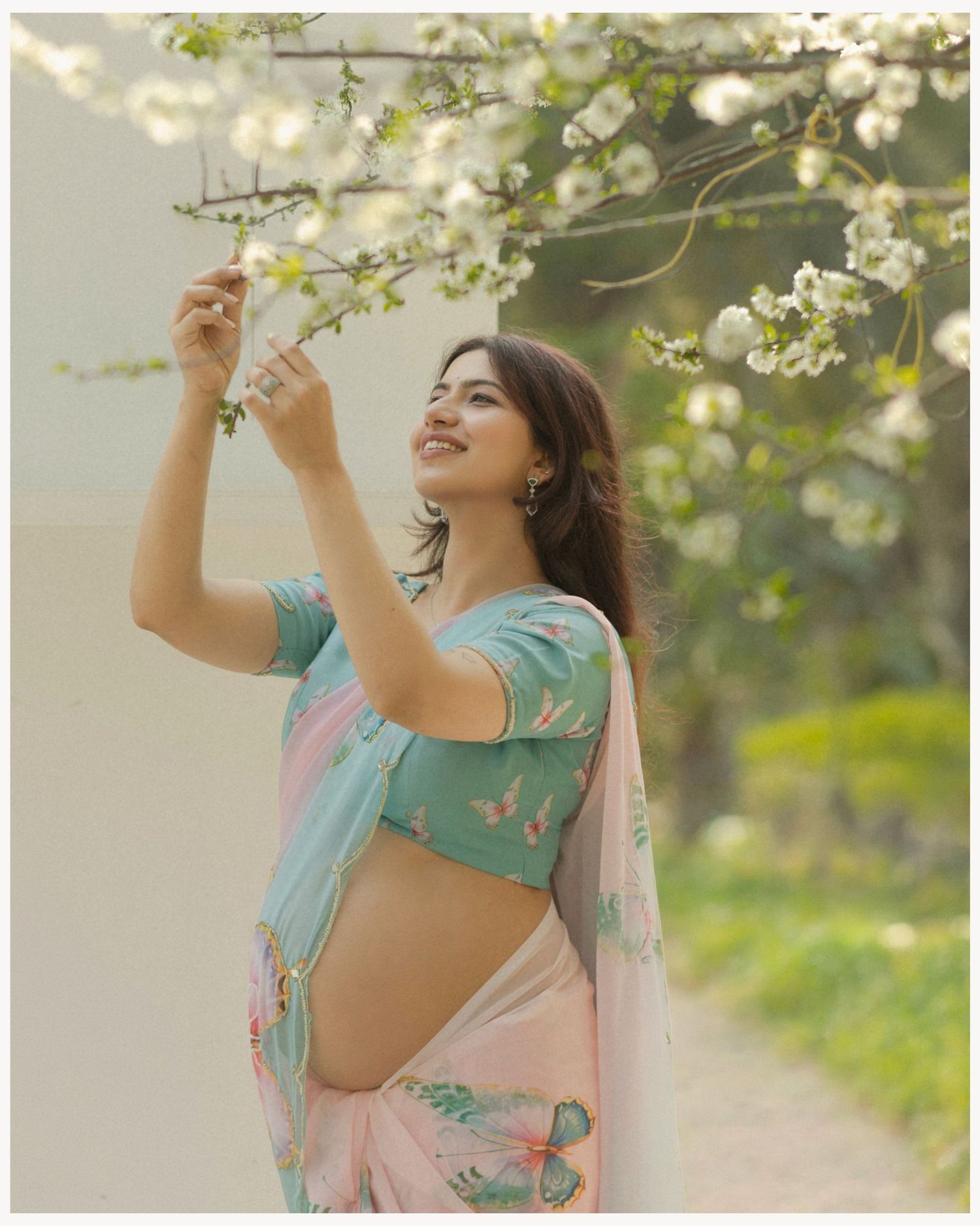 Woman in pink floral saree and teal blouse reaching for white blossoms on a tree branch outdoors.