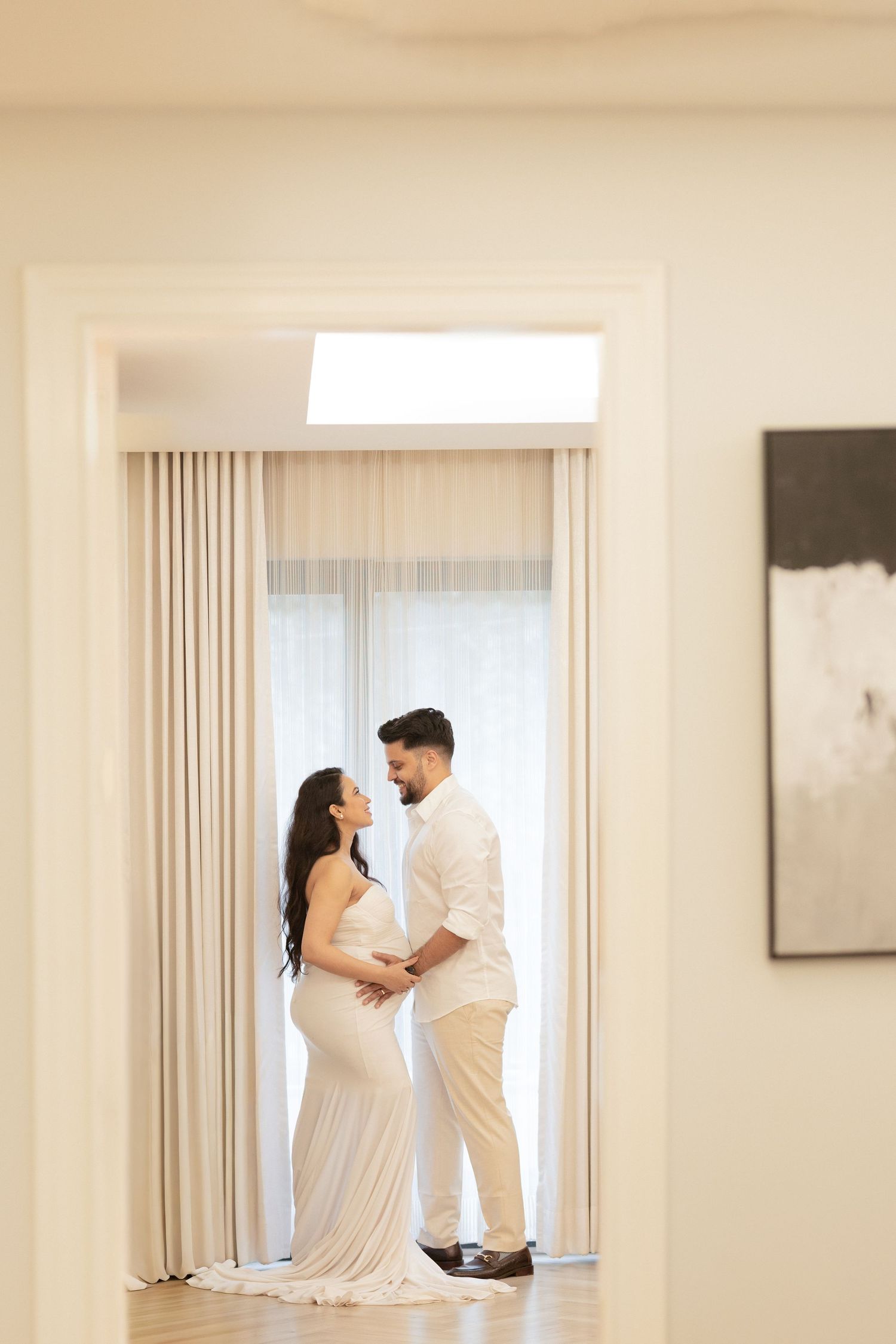 Couple in white outfits embracing near a bright window in an elegant room, woman in a fitted gown, man in dress shirt.