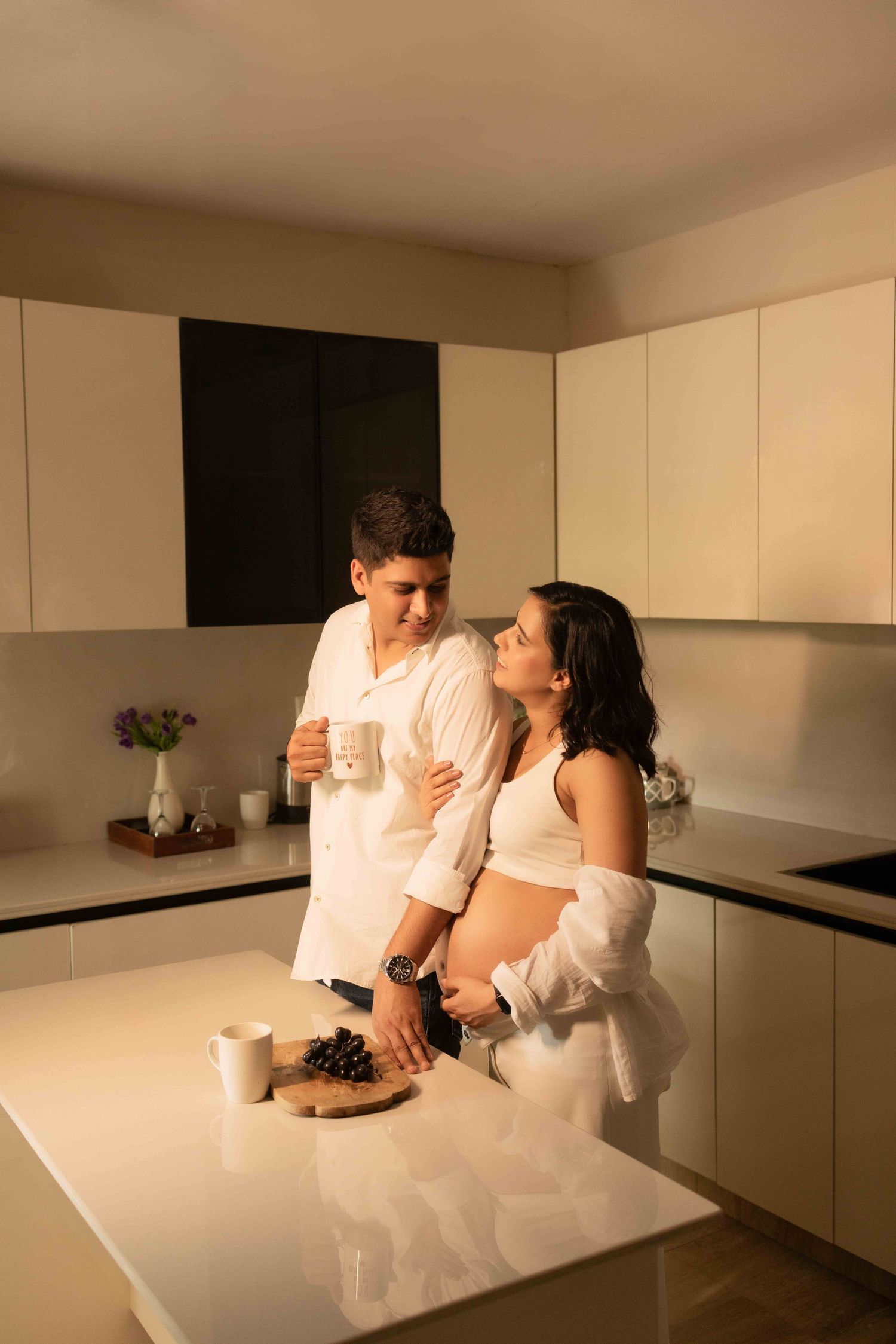 Pregnant couple sharing a tender moment in a modern white kitchen, man holding a mug while embracing his partner.