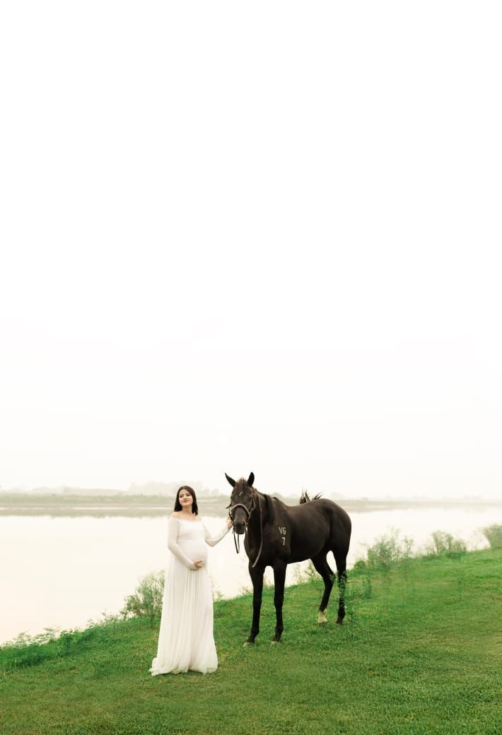 White-gowned figure stands with black horse against waterfront landscape.