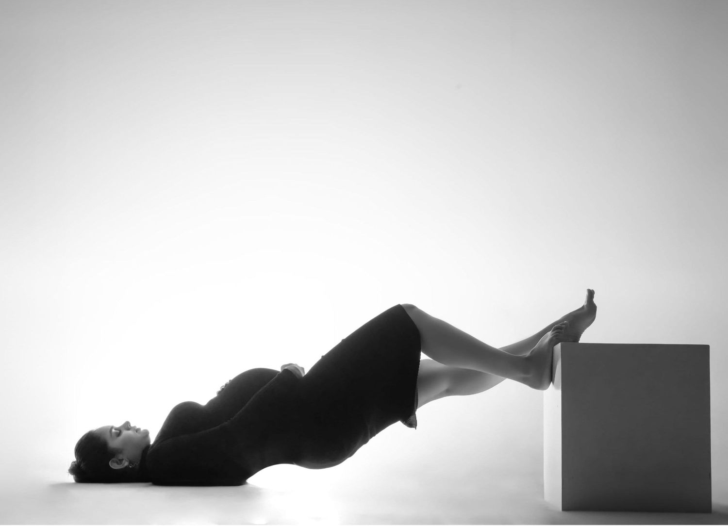 Black and white photo of a person lying on their back with legs resting on a white cube in a minimalist studio.