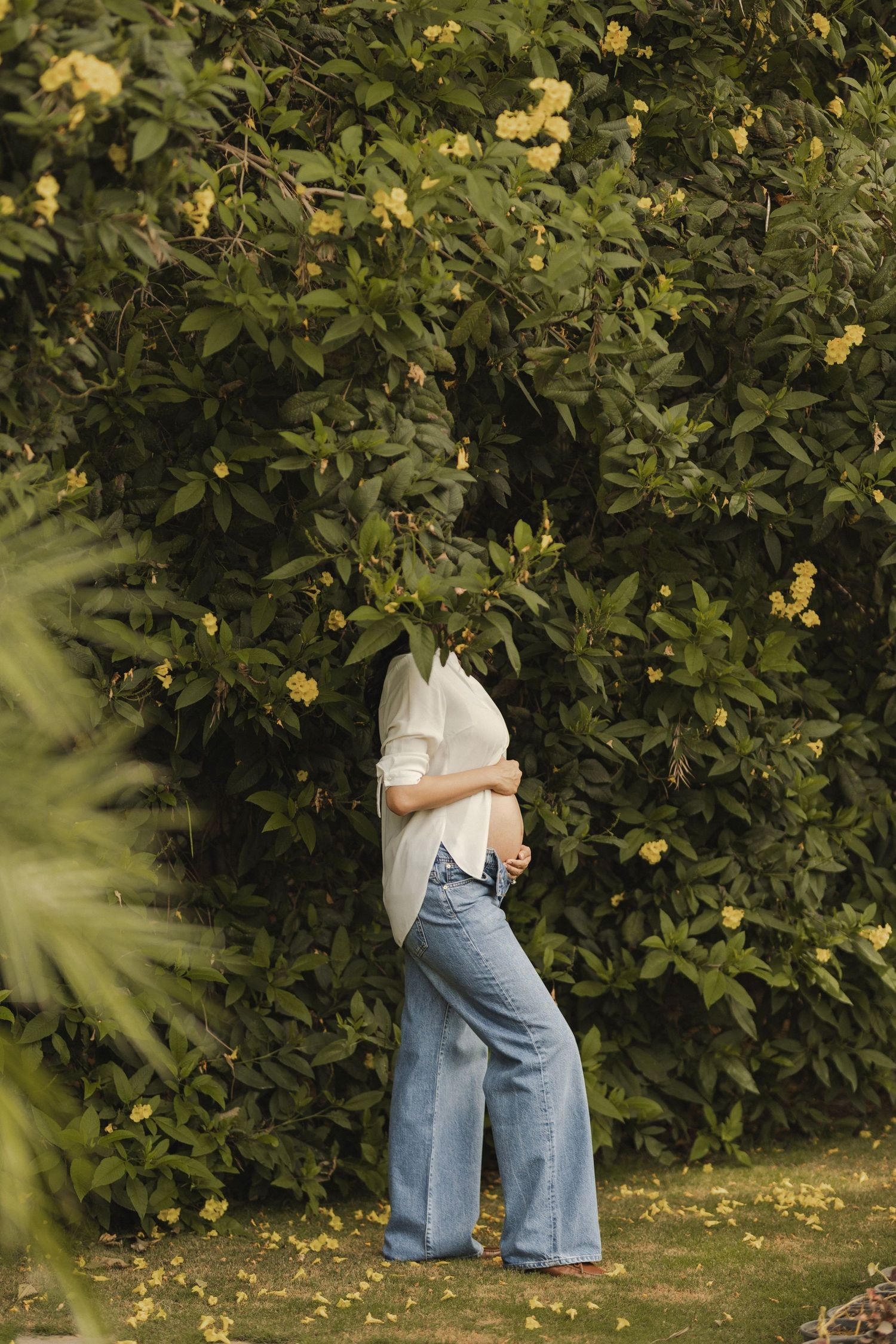 Pregnant woman in white shirt and wide-leg jeans cradling her baby bump near lush green flowering bushes outdoors.