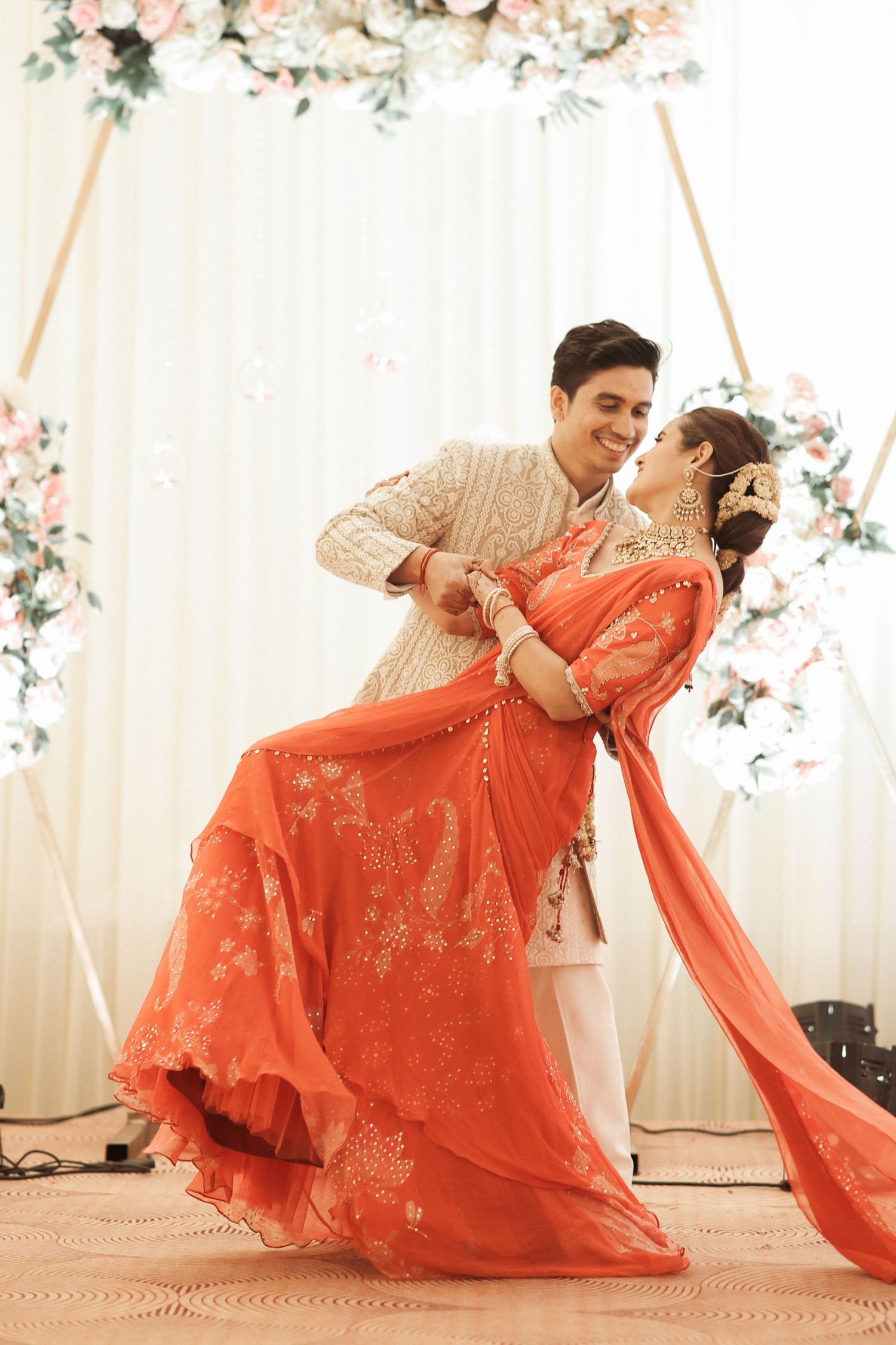 Couple dancing at Indian wedding, bride in orange embroidered lehenga, groom in cream sherwani, floral backdrop.