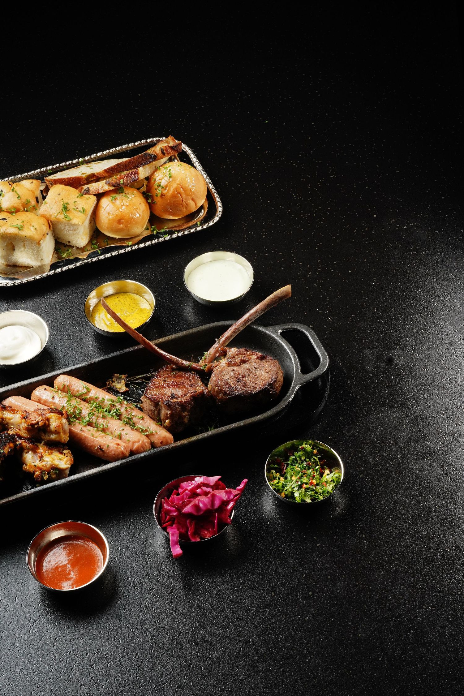 Assorted grilled meats and rolls served with dipping sauces and condiments on dark platters.
