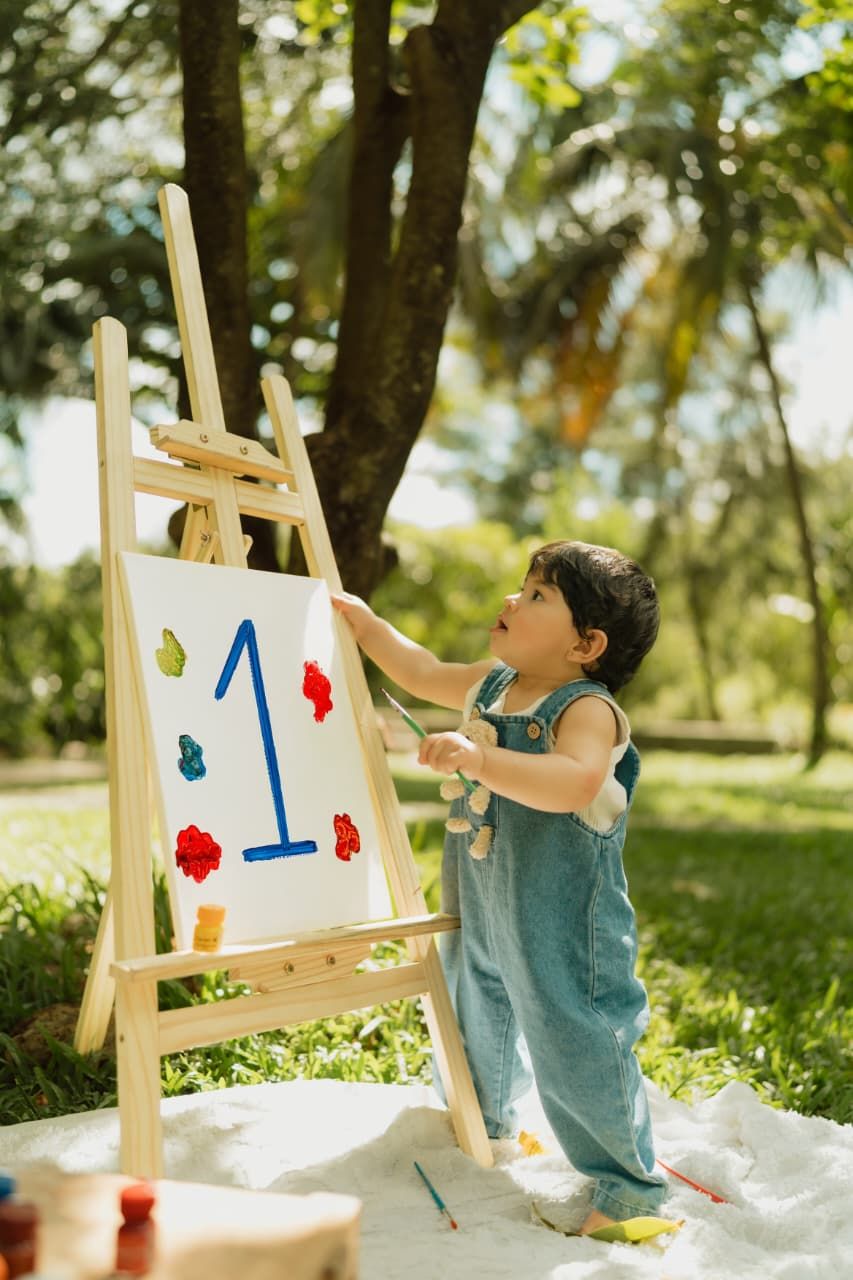 Toddler in denim overalls painting number 1 on easel outdoors, celebrating first birthday in green park.