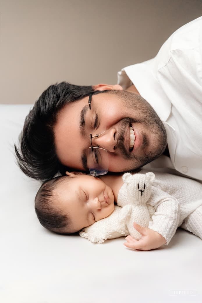 Smiling father with glasses lies beside sleeping newborn baby holding a white teddy bear on a white surface.