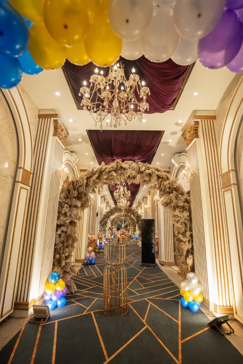 Luxurious event hall decorated with colorful balloons, floral arch, chandelier, and gold geometric carpet design.