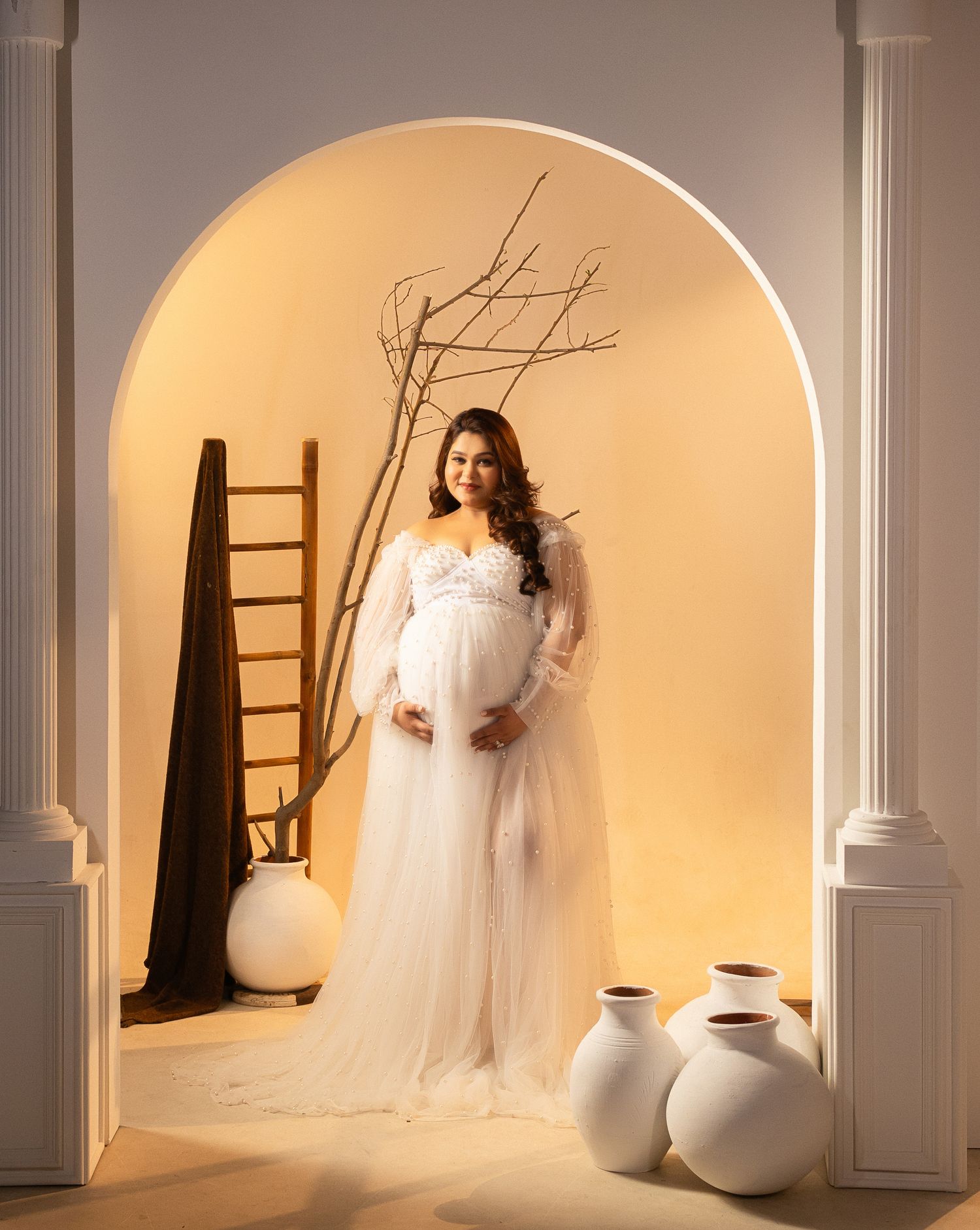 Elegant maternity photos in arched room with white gown and decorative vases.