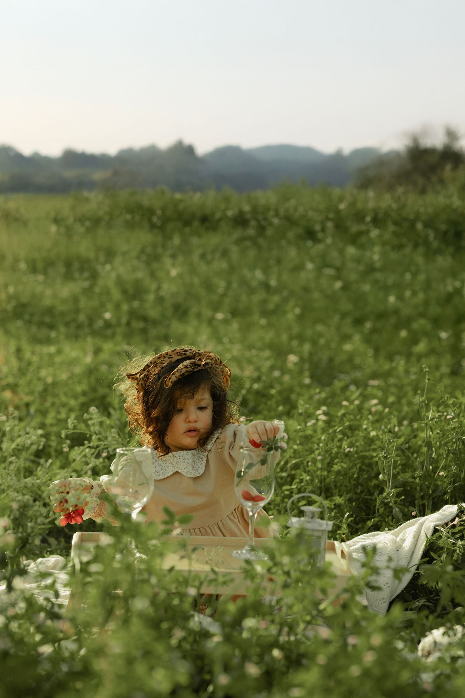 Young girl in floral dress and flower crown sits in lush green meadow holding a glass during a picnic.