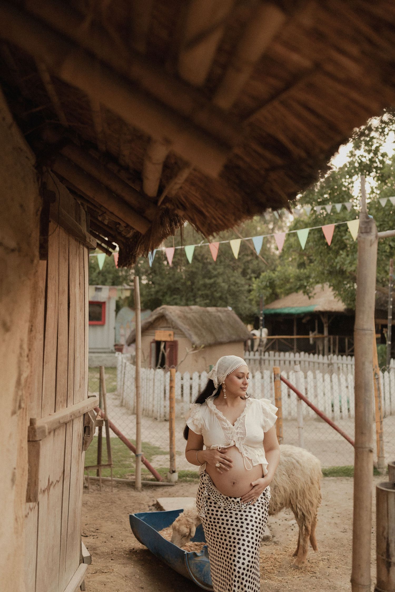 Pregnant woman in white crop top and polka dot skirt cradling her belly on a rustic farm with thatched roof.