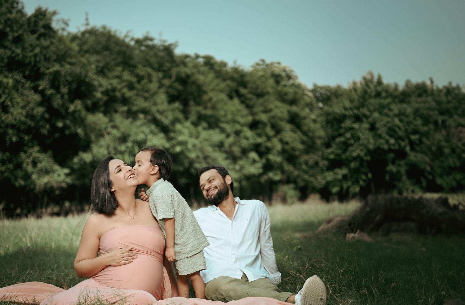 A family relaxes together on grassy field with trees in background during golden hour for a maternity photography.