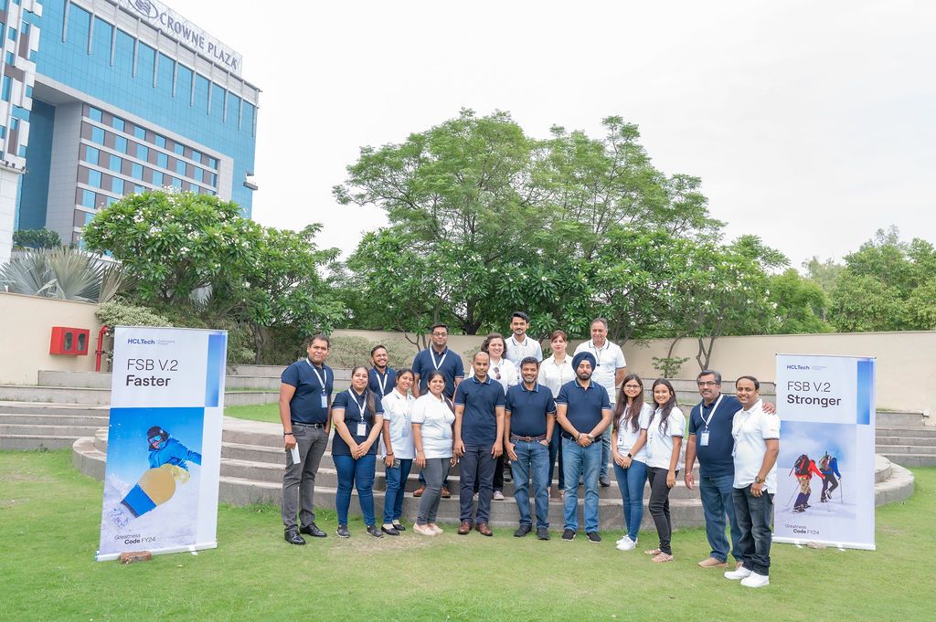 A diverse group of professionals pose outdoors near branded banners at a corporate team event in a modern business park.