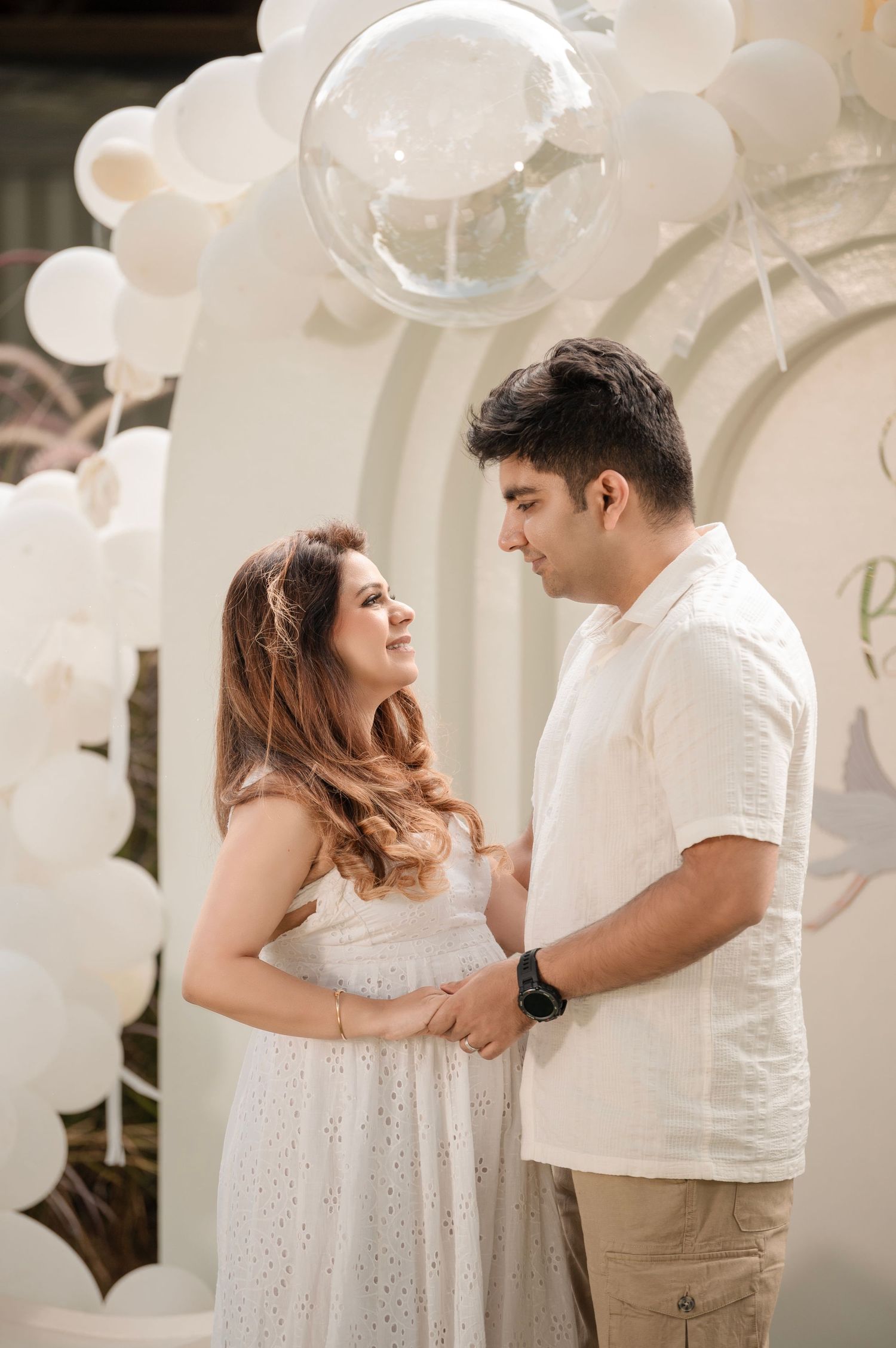 A couple in white outfits share a tender moment under a white balloon arch at a maternity photoshoot.