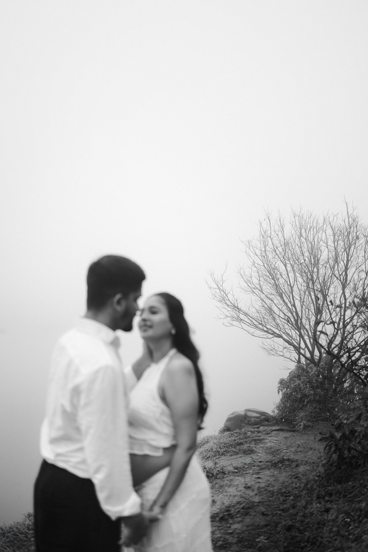 A romantic black and white photograph of a couple embracing on a misty hilltop with bare branches in the background.