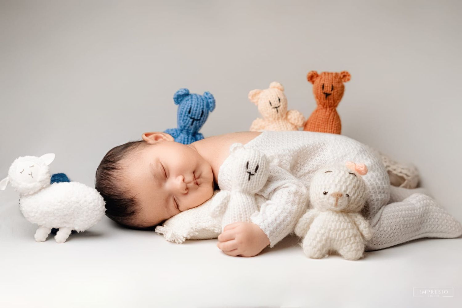 Sleeping newborn baby surrounded by colorful knitted stuffed animals including bears and a lamb on white background.
