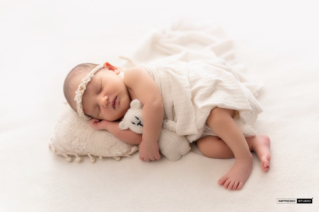 Sleeping newborn baby in white dress with headband resting on pillow hugging small teddy bear on white background.