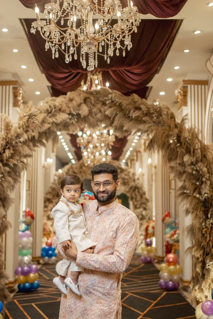 Man in traditional kurta holds toddler in decorated wedding hall with pampas grass arches and chandeliers.