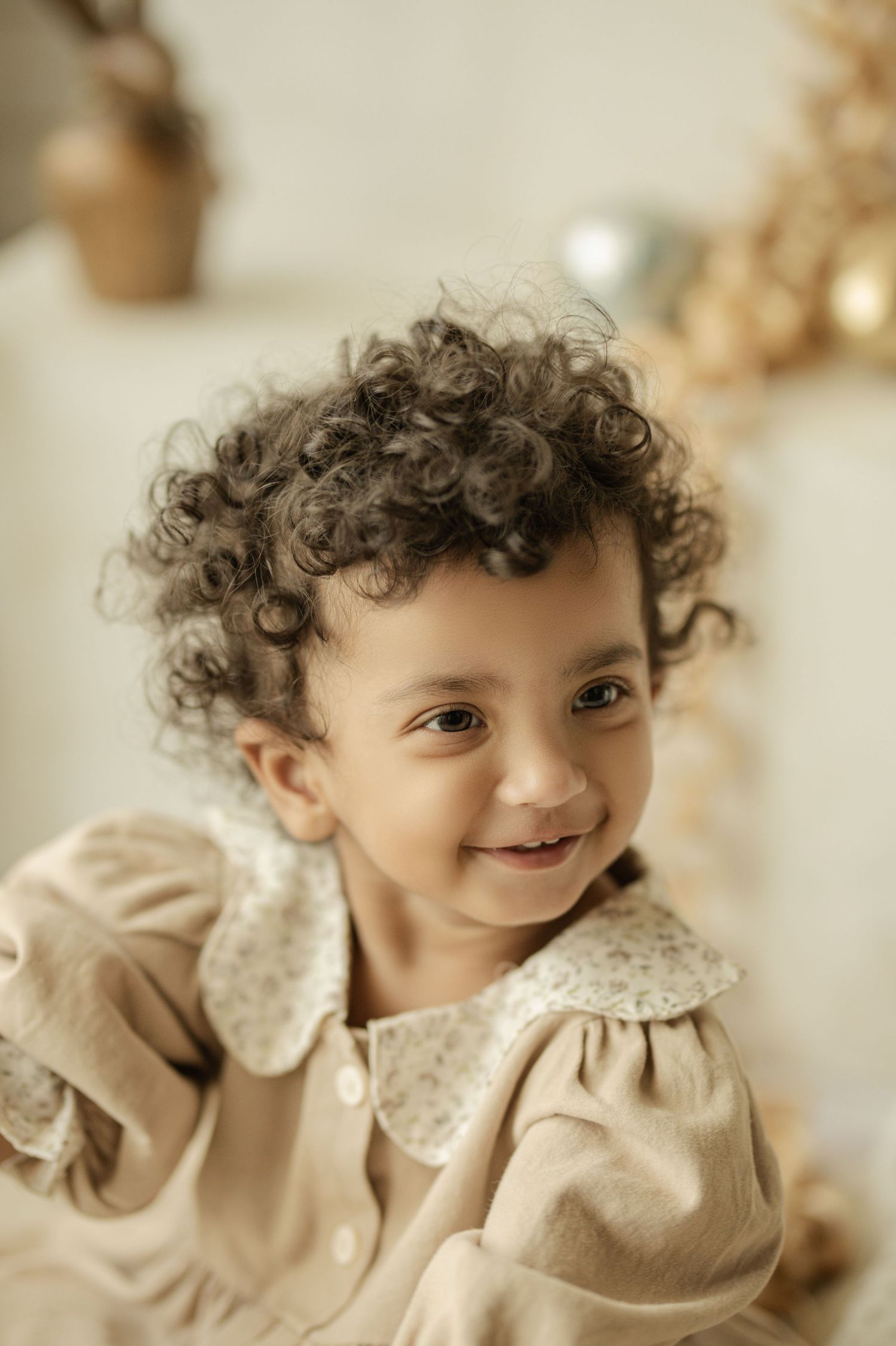 Adorable toddler with curly hair smiling, wearing a beige floral collar dress in a soft, neutral-toned setting.