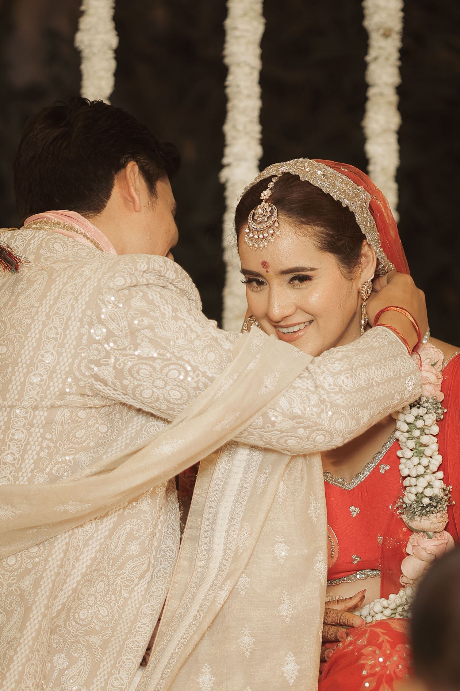 Bride in red lehenga and groom in white sherwani share a tender moment during traditional Indian wedding ceremony.