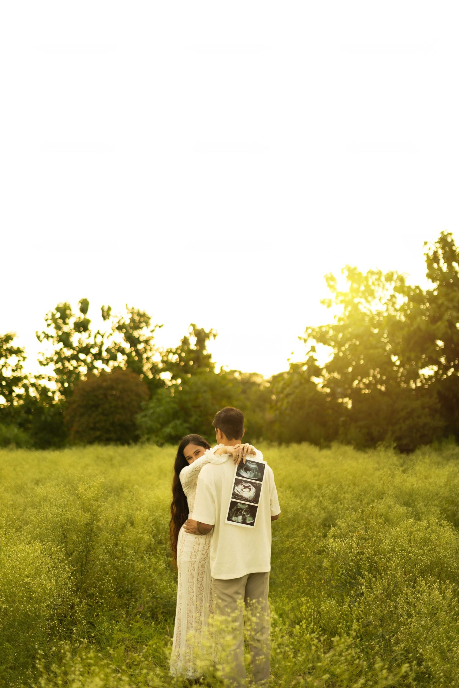 Couple embracing in a sunlit green field, man holding ultrasound photos on his back, announcing pregnancy at golden hour.