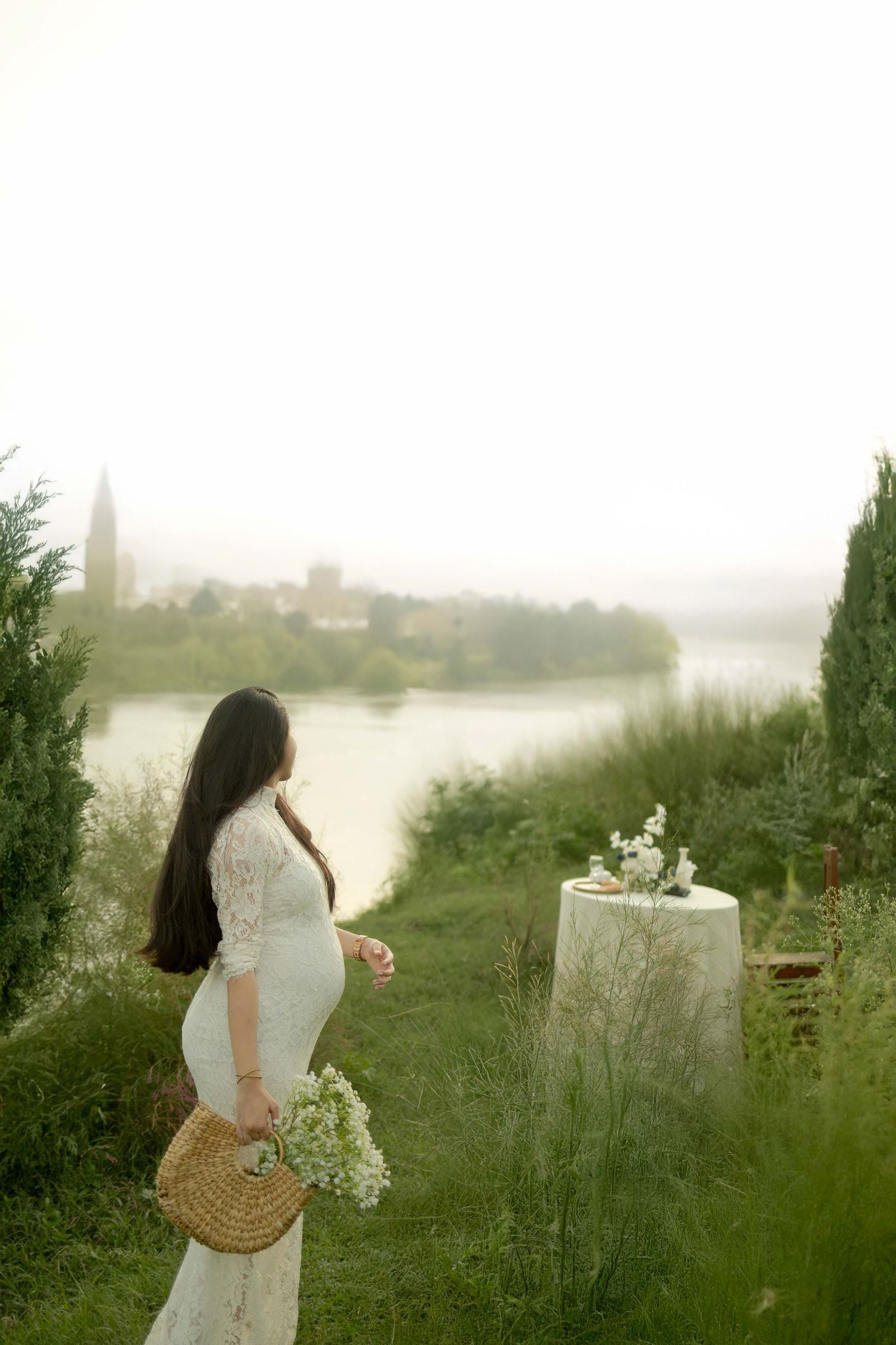 Woman in white lace dress holding wicker basket with flowers, standing by misty riverside garden.
