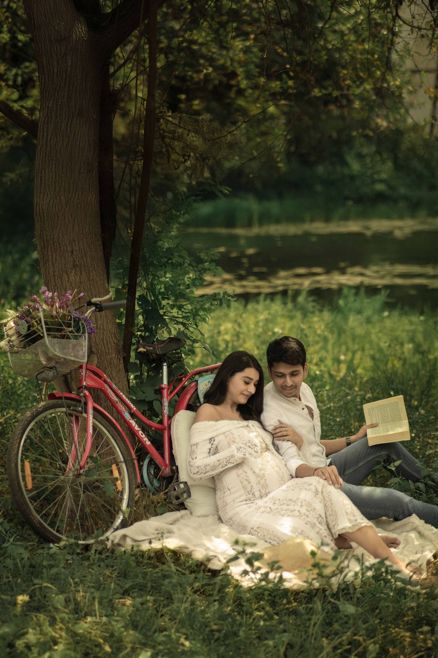Romantic couple reading a book together on a blanket under a tree near a lake, with a vintage pink bicycle nearby.