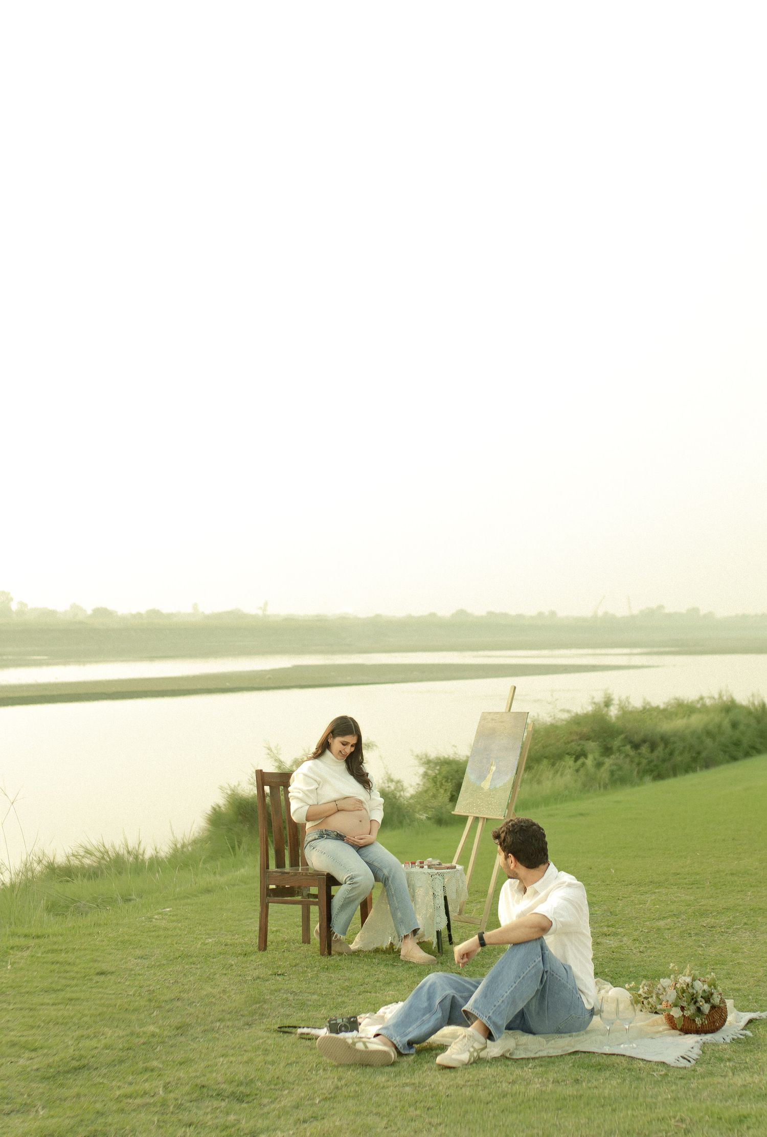 A couple enjoys a peaceful outdoor maternity photoshoot by a lake, with an easel, chair, and picnic blanket on green grass.