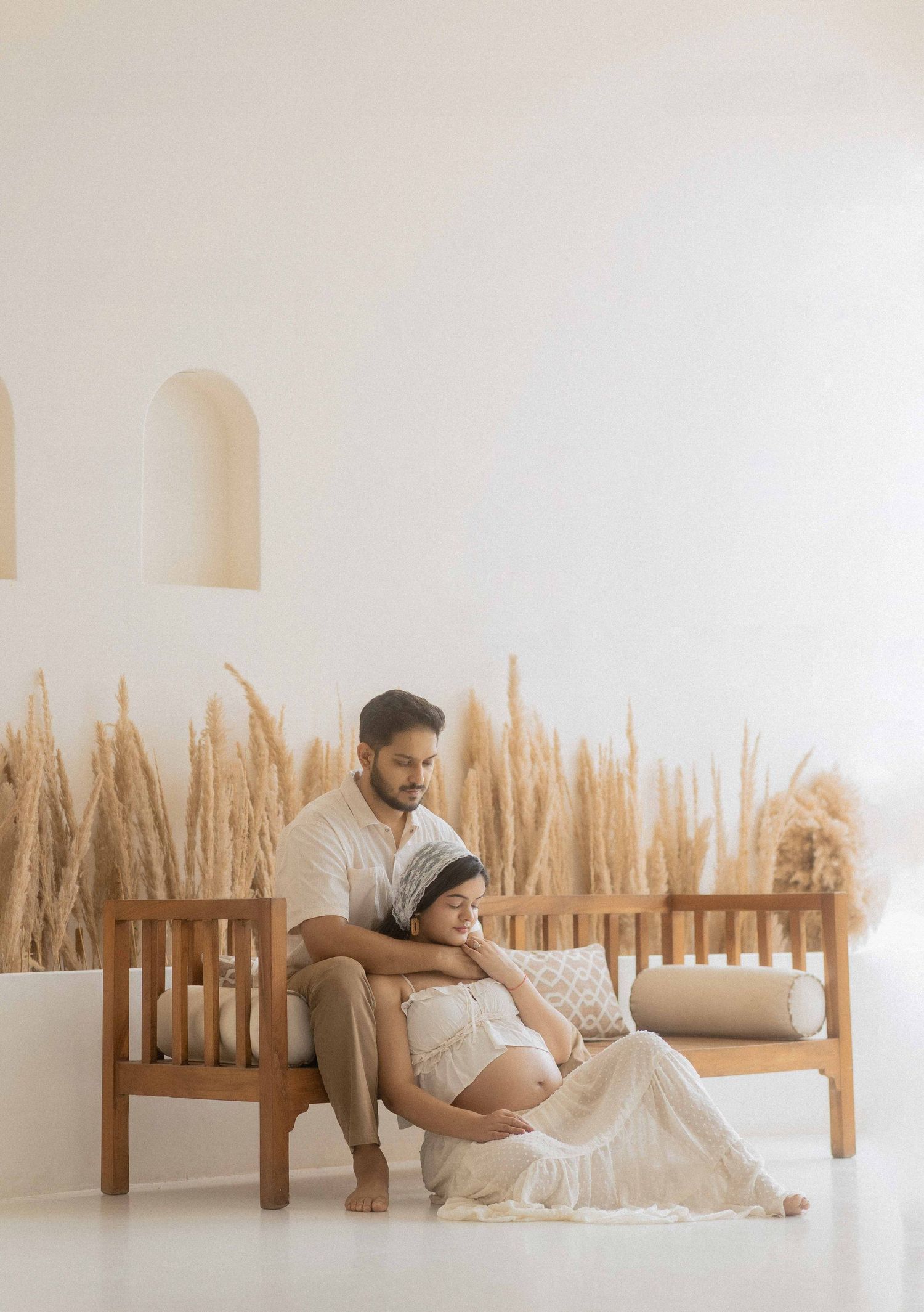 Couple in boho maternity photoshoot, pregnant woman in white dress sits on floor leaning on man seated on wooden bench.