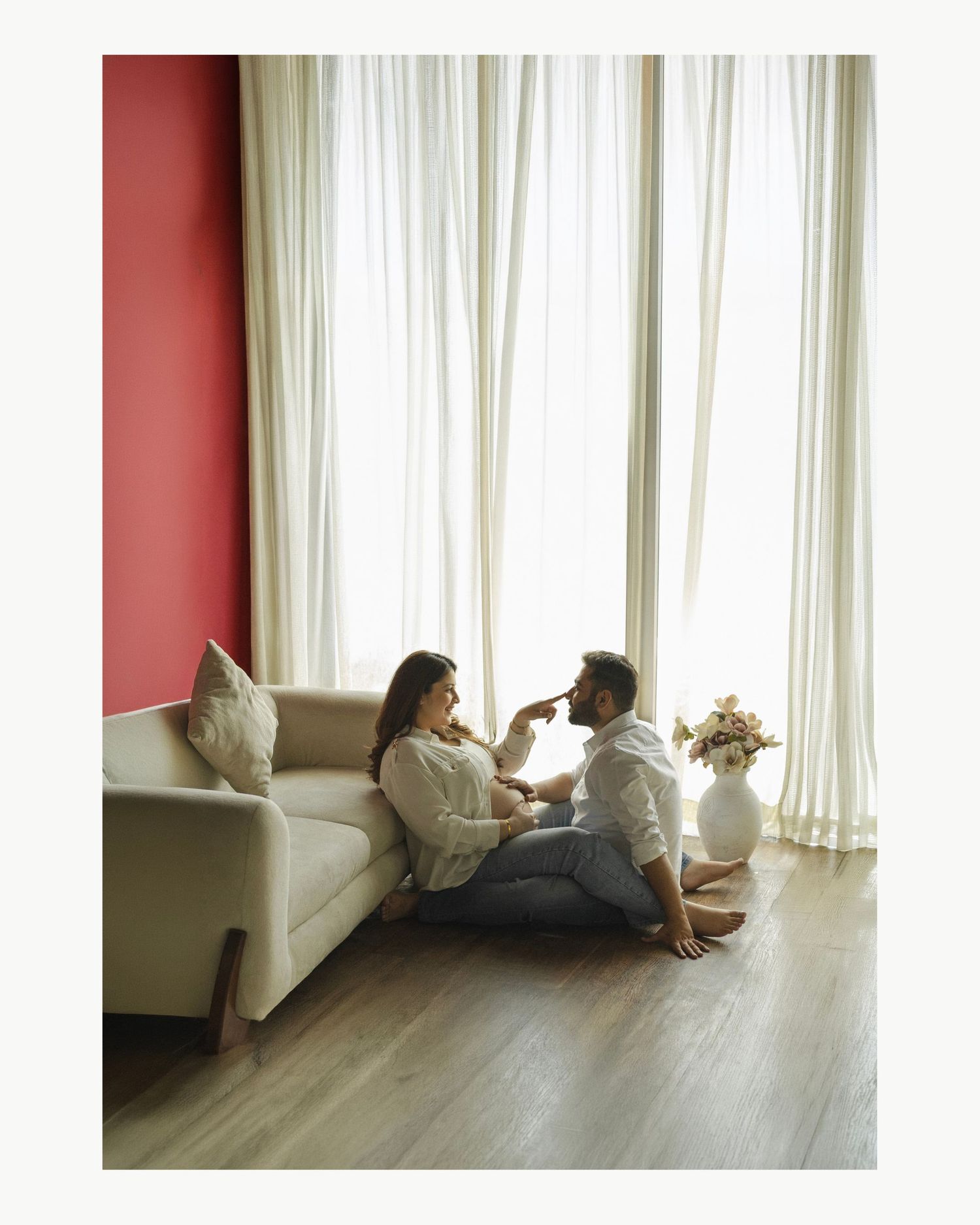 Couple sitting on floor by sofa, sharing a tender moment near large curtained windows in a modern living room.